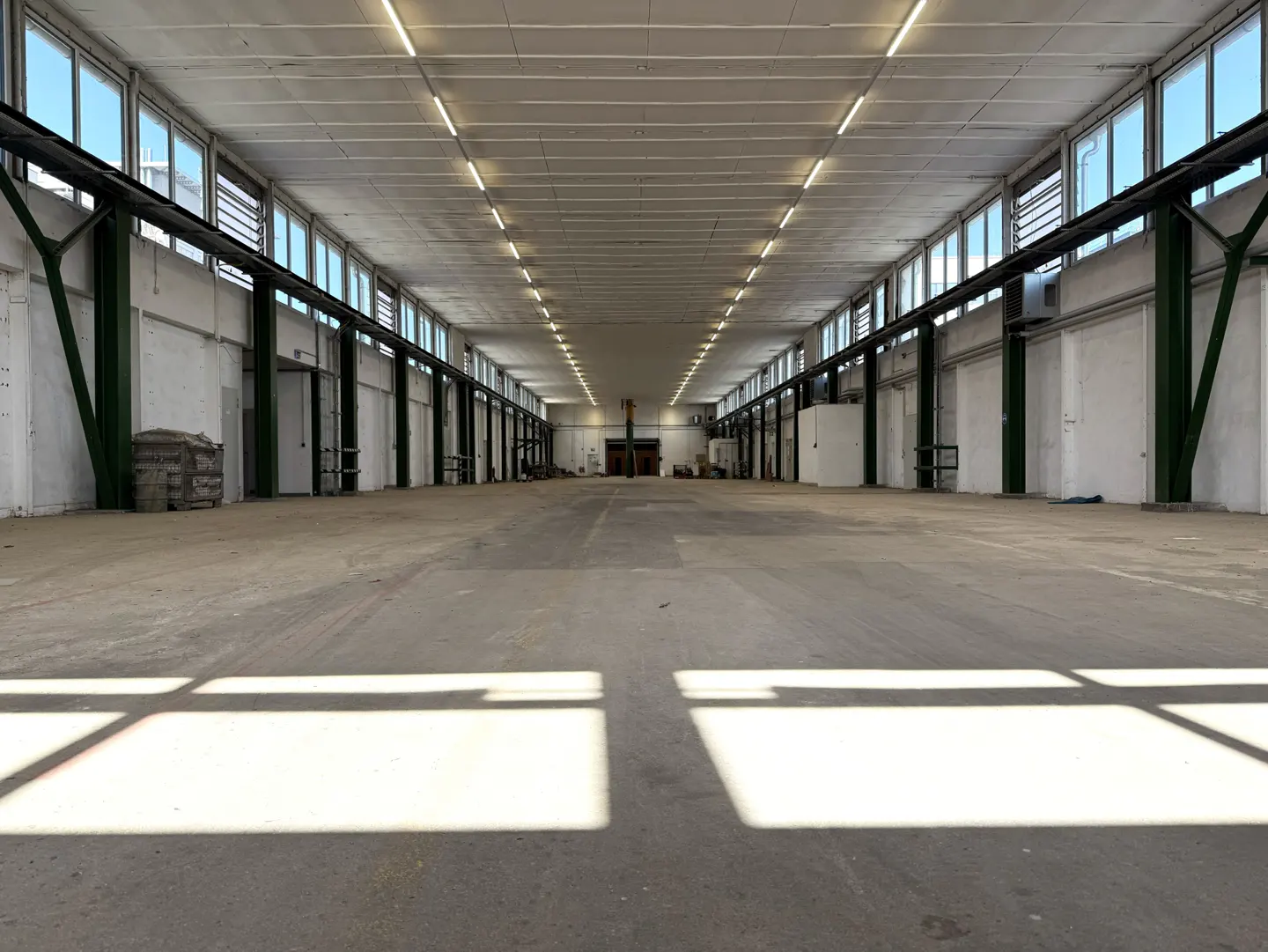 A large, empty warehouse with concrete floors, white walls, and rows of windows. Green support beams line the walls.