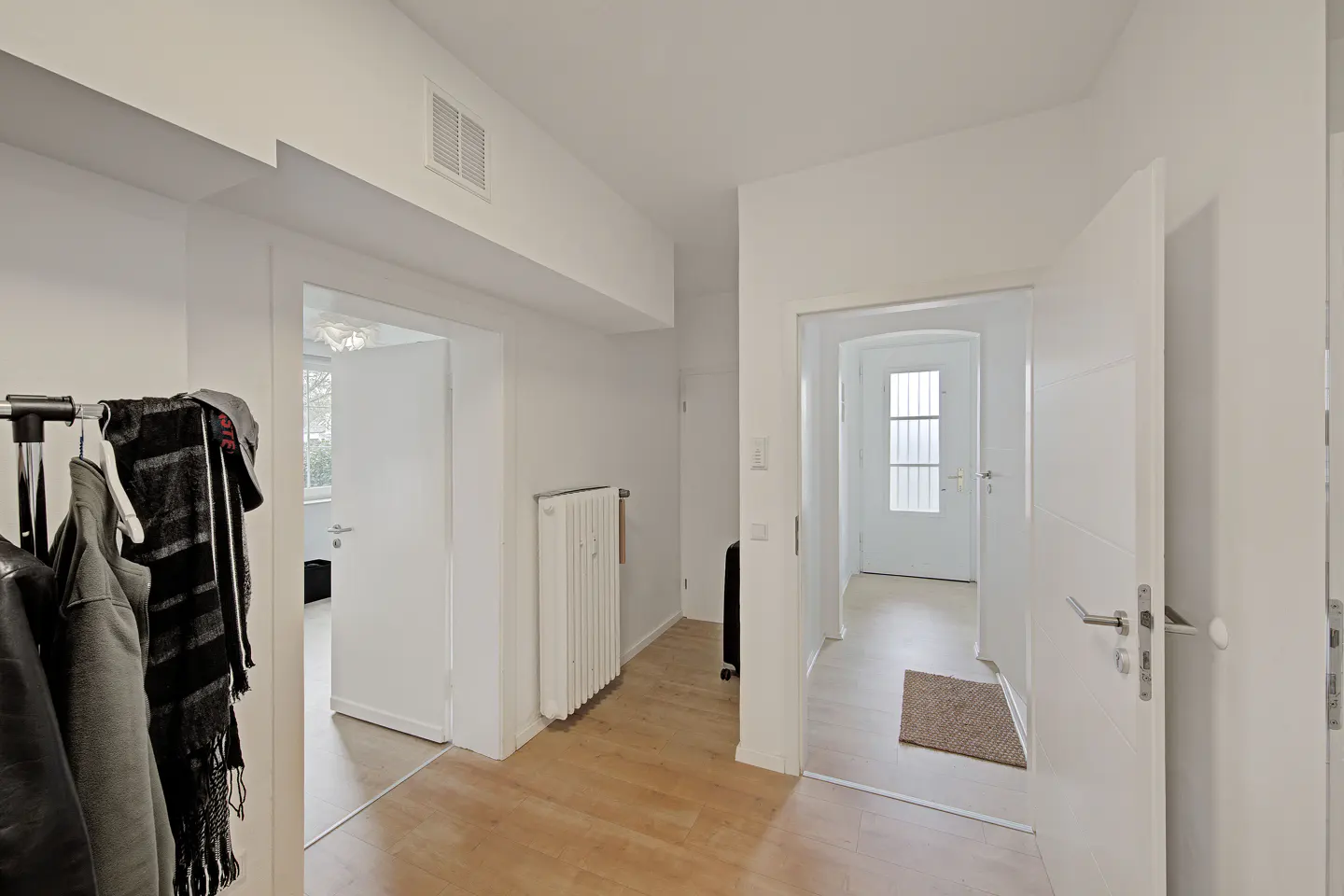 Bright, white hallway with light wood floors. A coat rack with clothes is on the left. Open doors lead to other rooms.