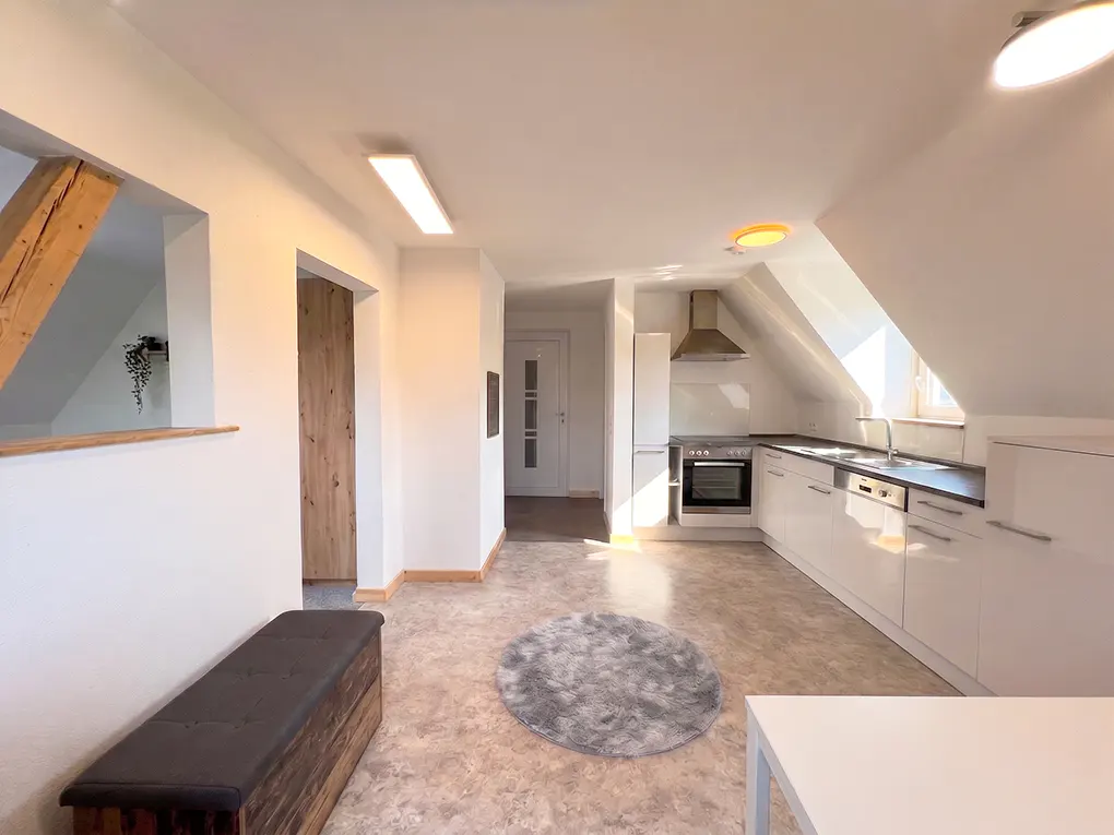 Bright, open-plan attic apartment with white walls, a modern kitchen, and a wooden bench. A gray rug sits on the floor.