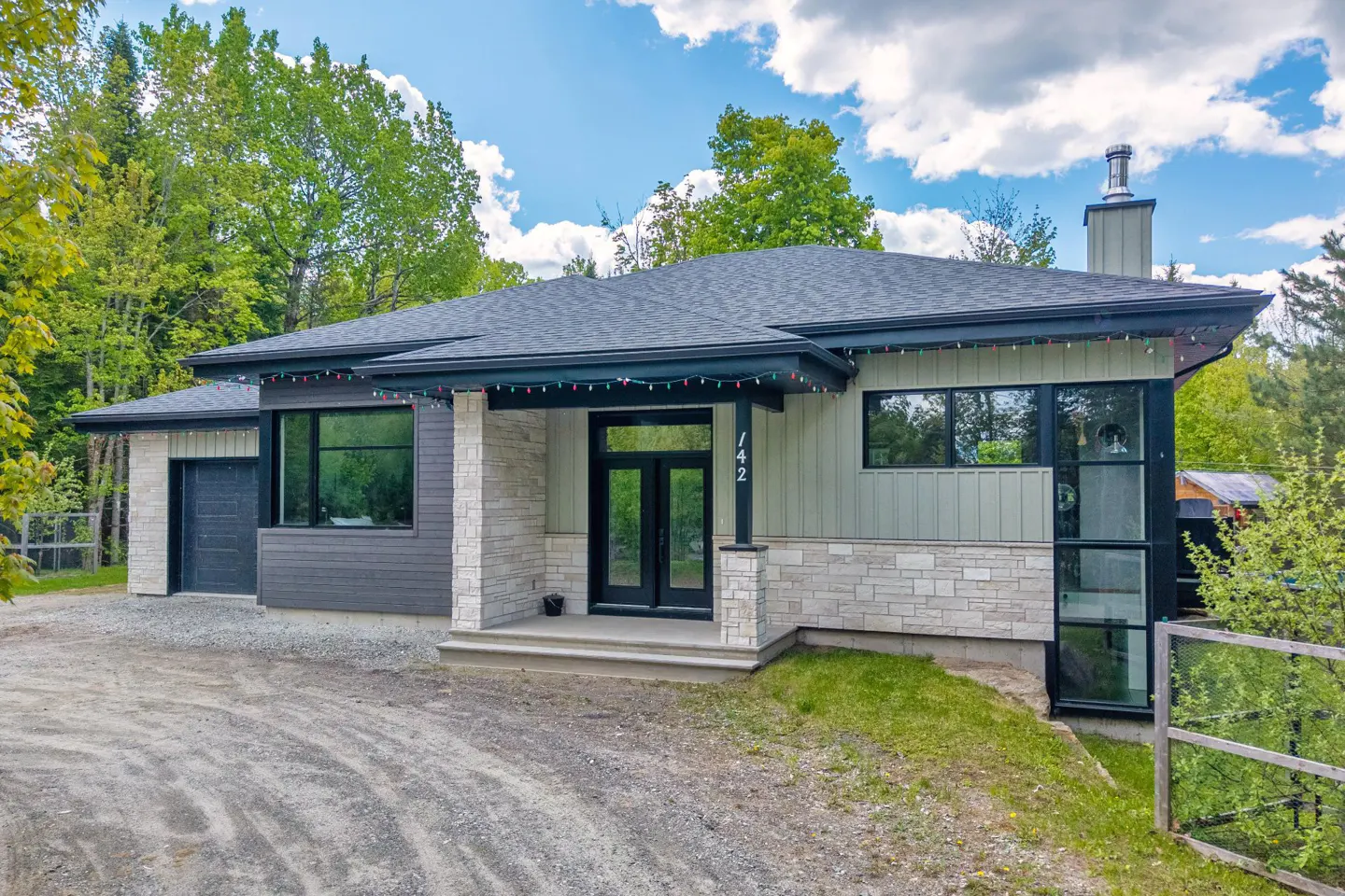 Modern single-story home with stone and light green siding, black trim, and a gravel driveway surrounded by lush trees.