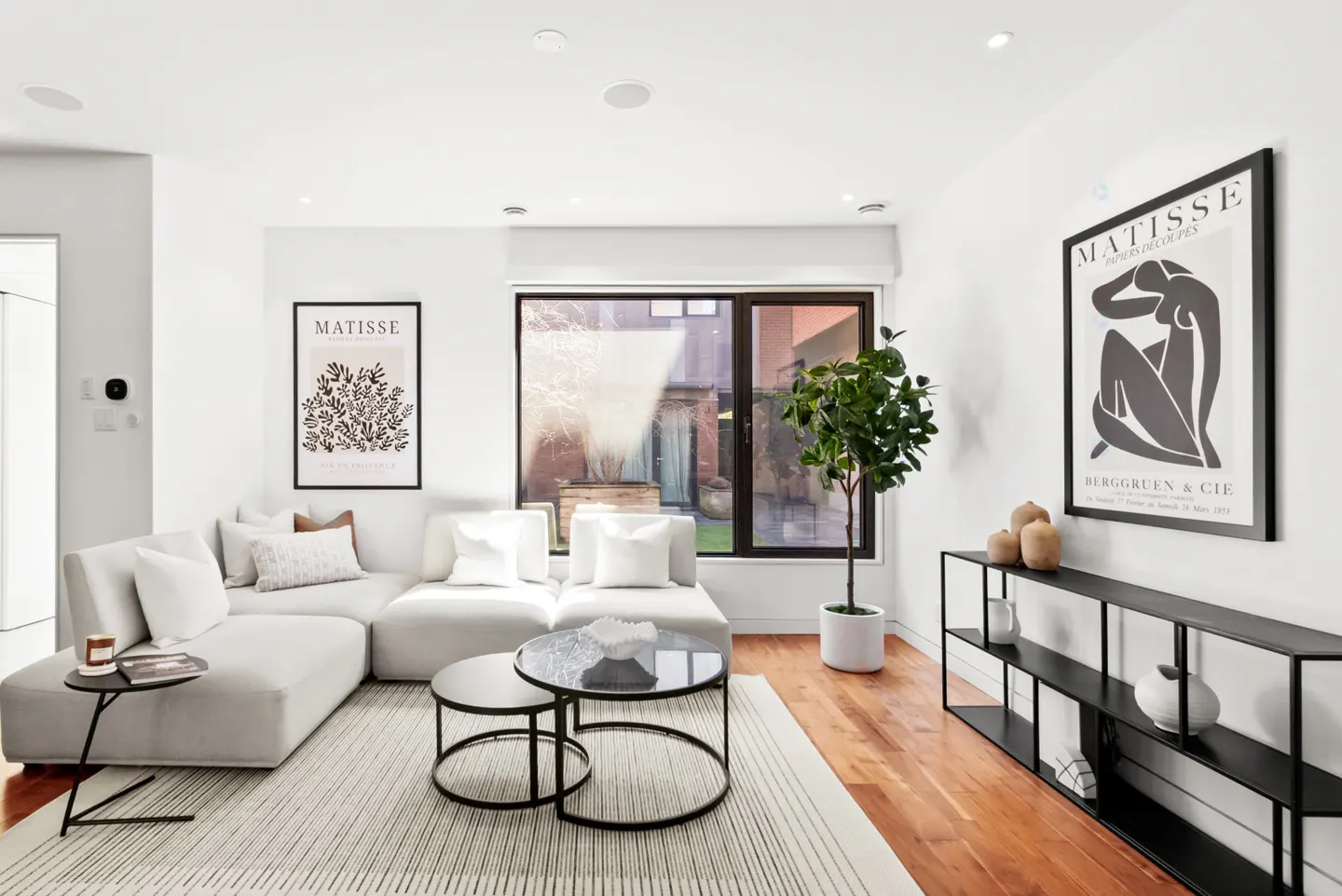 Bright living room with white walls, light gray sectional sofa, black tables, and Matisse art prints. Hardwood floors and a large window add warmth.