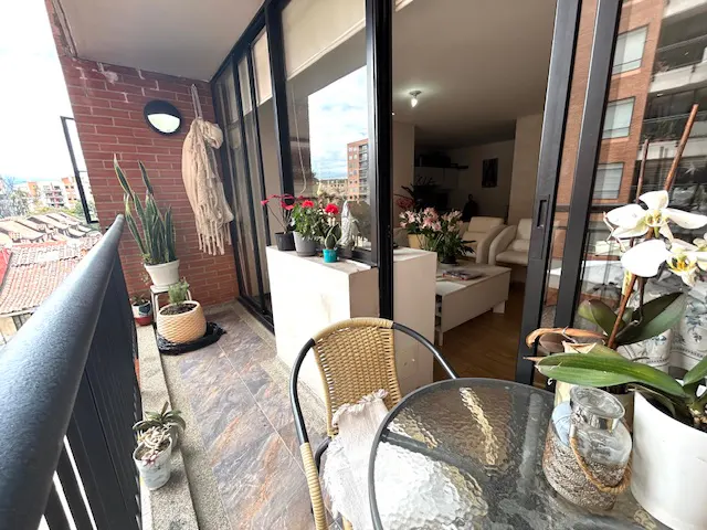 A balcony with a glass table, wicker chair, and potted plants overlooks a city. Sliding glass doors lead to a living room with white furniture.