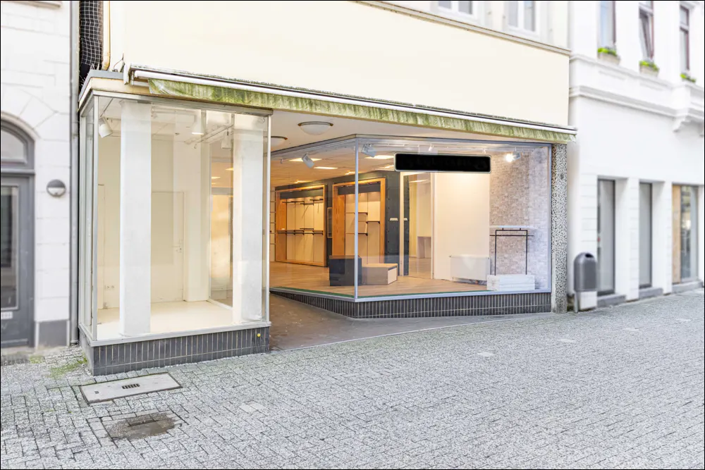 Storefront with glass windows and awning on a cobblestone street. Inside, racks and shelves are visible.