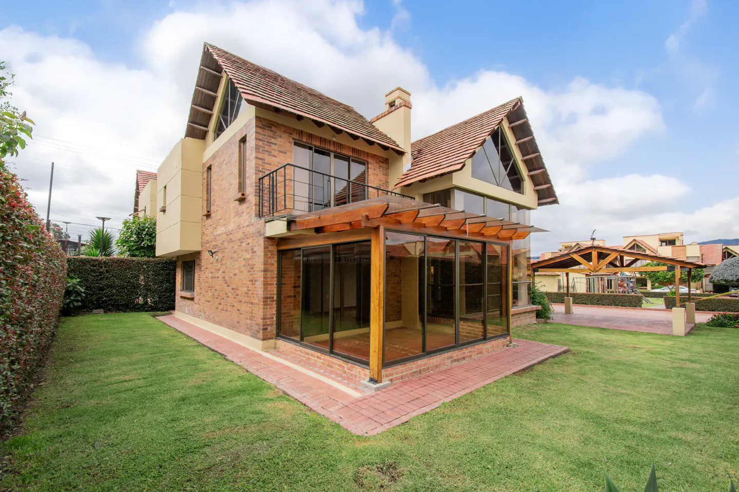 Two-story brick house with a glass-enclosed patio, wooden pergola, balcony, and a large green lawn under a cloudy sky.
