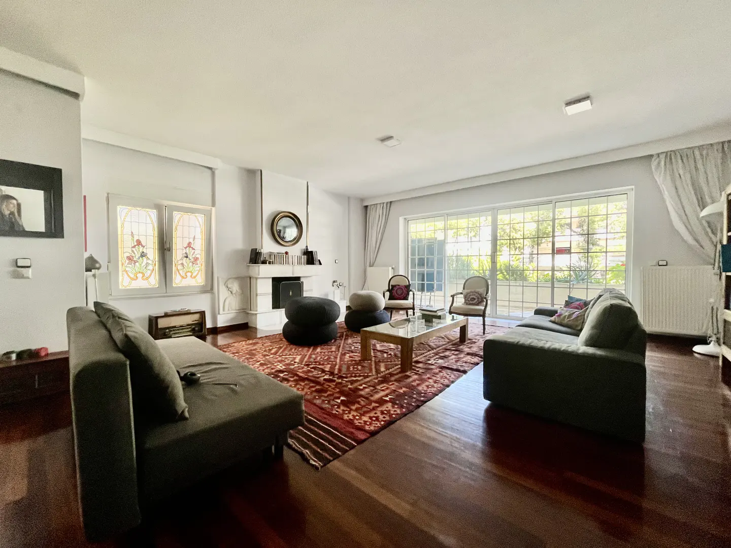 Bright living room with hardwood floors, a red patterned rug, and two gray sofas. A fireplace, stained glass windows, and a sliding glass door to the outside are visible.