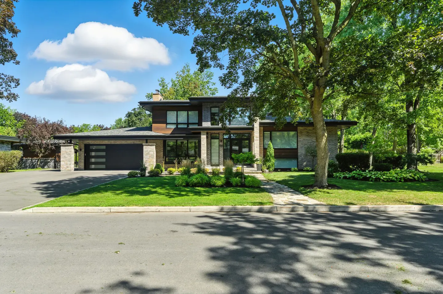 Modern two-story home with stone and wood facade, black garage door, green lawn, and mature trees under a blue sky.