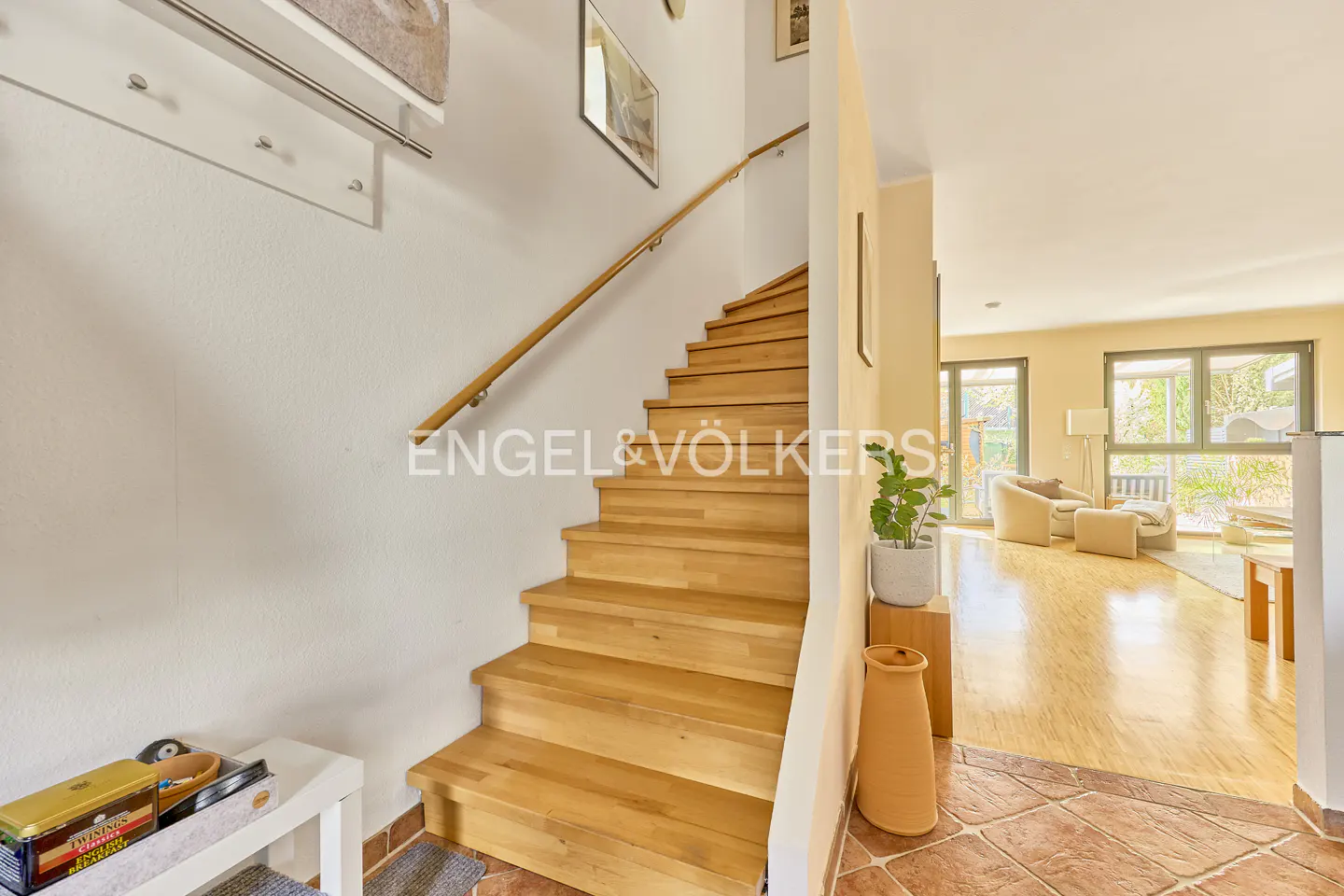 Interior view of a home featuring a wooden staircase with a light wood handrail, leading to a bright living room with large windows.