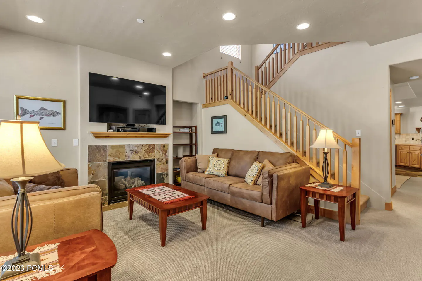 Living room with a stone fireplace, wall-mounted TV, brown leather sofa, wooden coffee table, and stairs with wood railing.