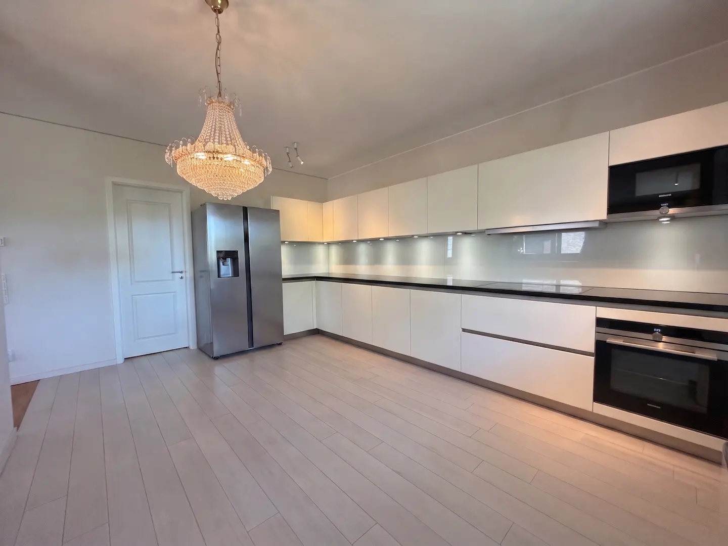 Bright kitchen with white cabinets, black countertops, and stainless steel appliances. A crystal chandelier hangs from the ceiling.