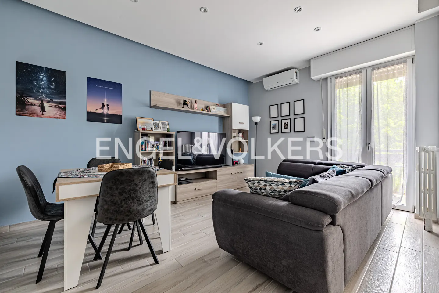 Living room with a dining table, gray sofa, TV, and light blue walls. Artwork hangs on the wall. Natural light filters through sheer curtains.
