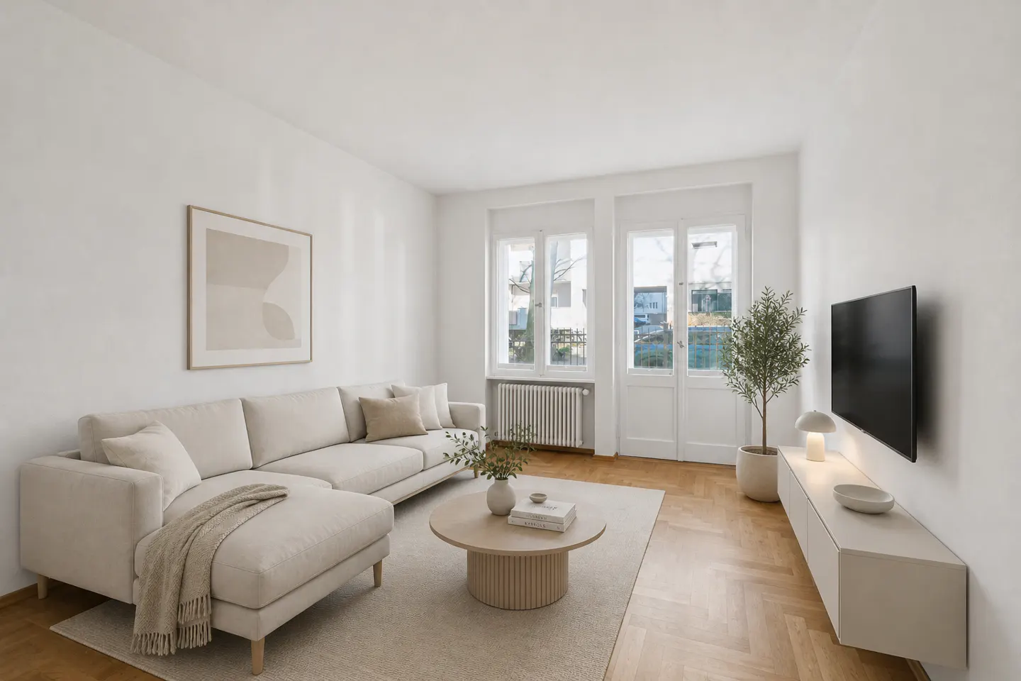 Bright living room with white walls, herringbone wood floors, and neutral-toned furniture. A sectional sofa faces a TV, with a round coffee table in between.