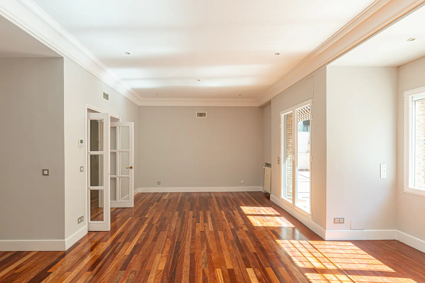 Bright, empty room with hardwood floors, gray walls, and white trim. French doors and windows let in sunlight.