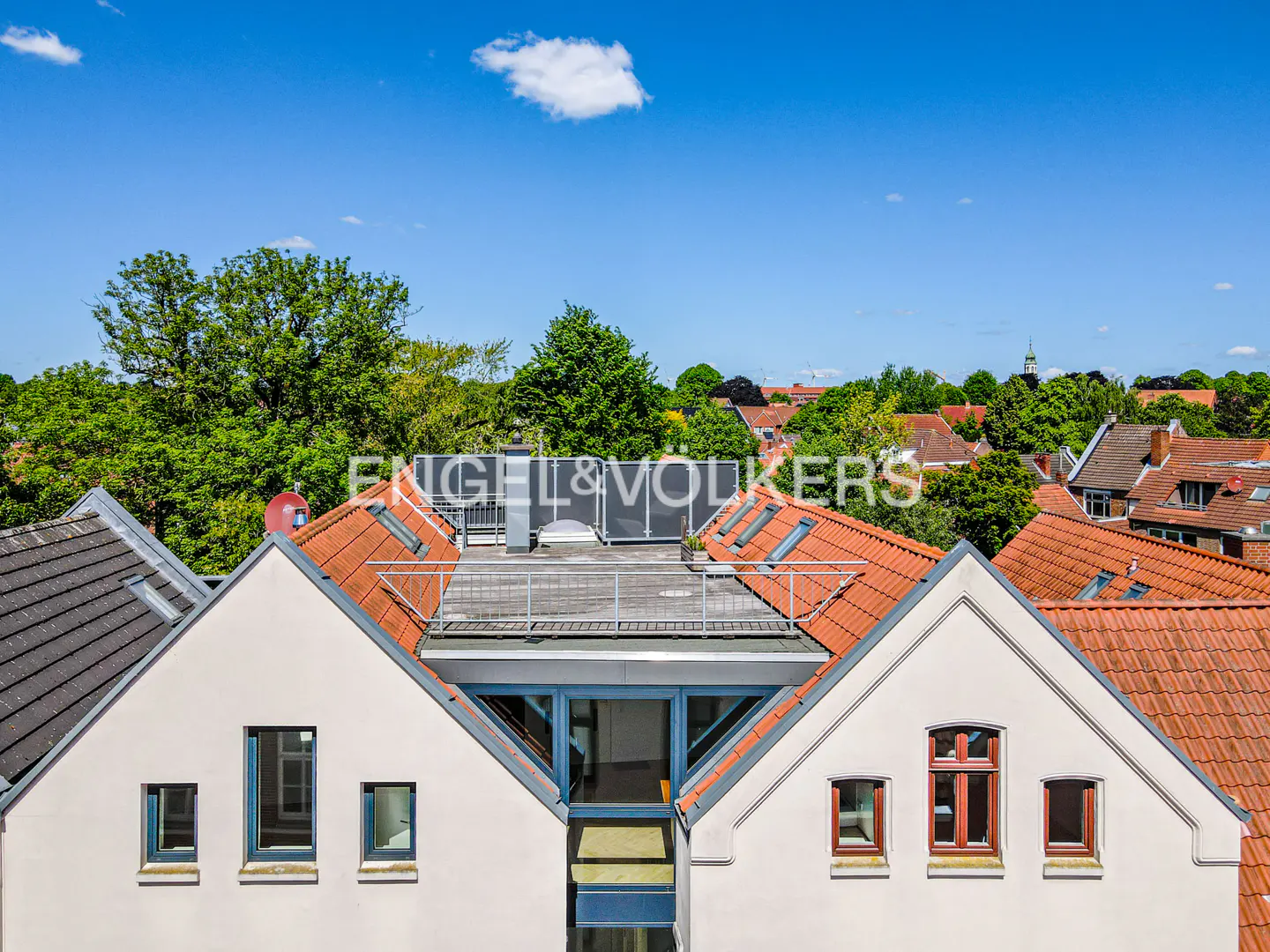 View of rooftops with red tiles and white walls under a blue sky. The Engel & Völkers logo is visible in the background.