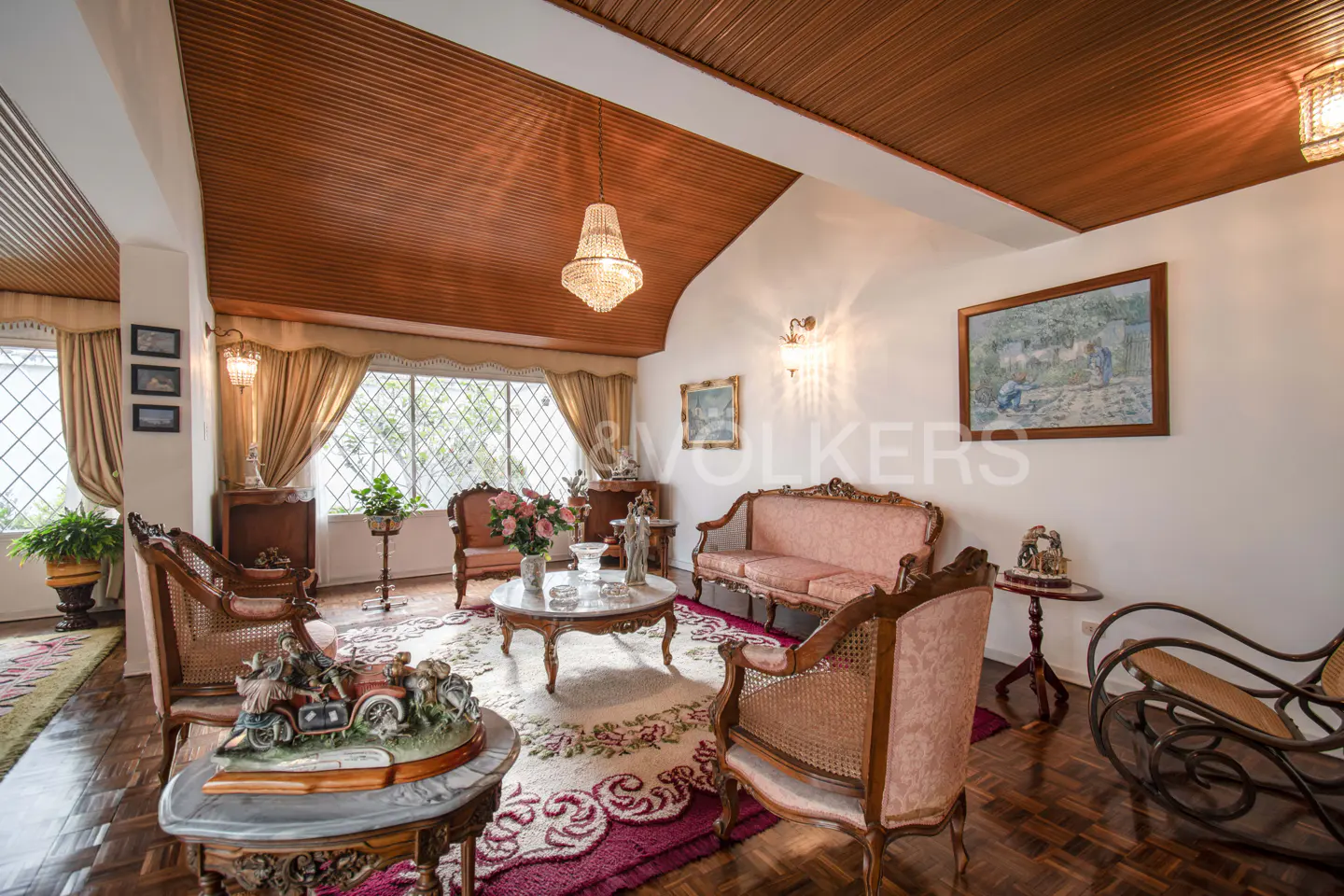 Living room with wood floors, pink sofas, and ornate chairs. A chandelier hangs from the wood-paneled ceiling. Large windows with gold curtains.