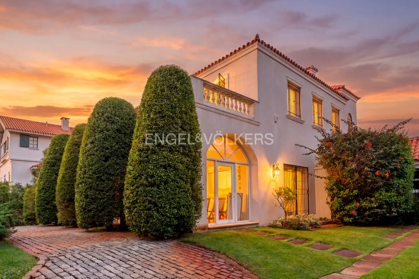 Exterior of a white two-story house with a red tile roof at sunset. Tall, green bushes line the brick driveway.