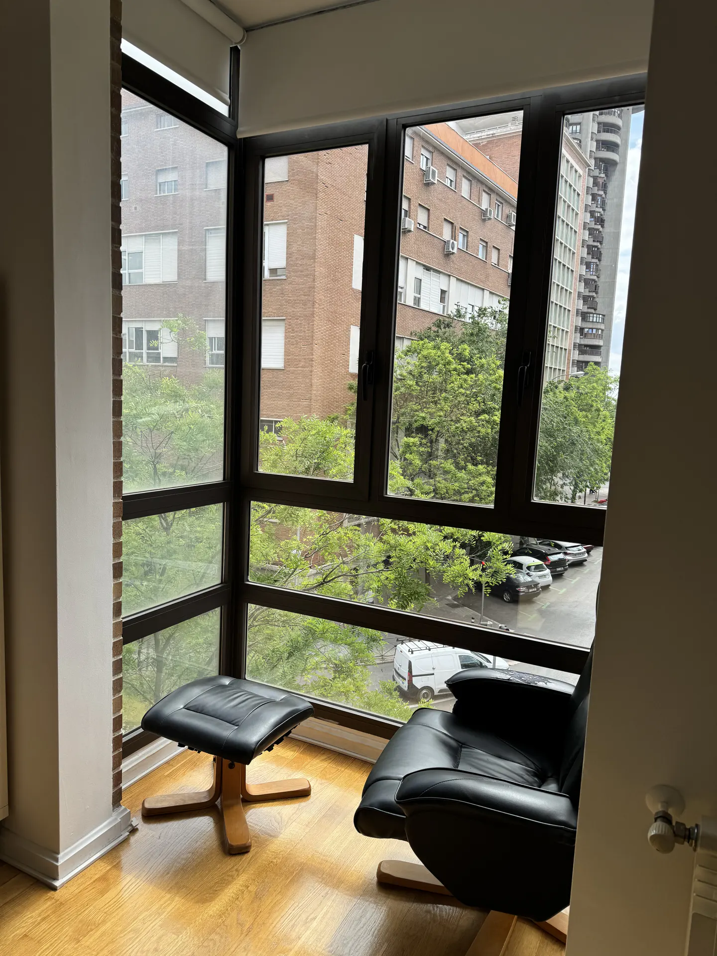 Corner view of a room with a black leather chair and ottoman, a large window with a city view, and hardwood floors.