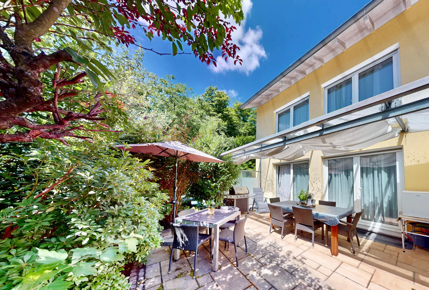 A sunny patio with two tables and chairs, a grill, and lush greenery next to a yellow house.
