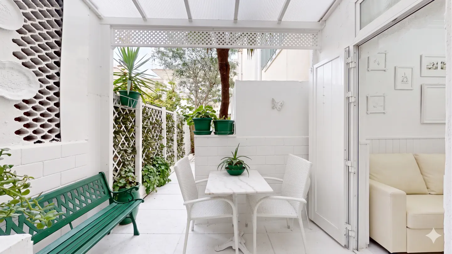 Bright, all-white patio with a green bench, white table and chairs, and potted plants. A white door leads to a cream-colored sofa.