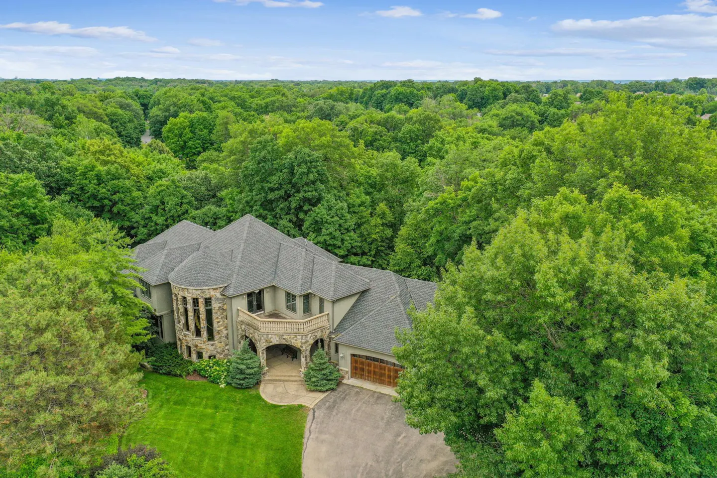 Aerial view of a large, stone and stucco house with a gray roof, surrounded by lush green trees and a blue sky.