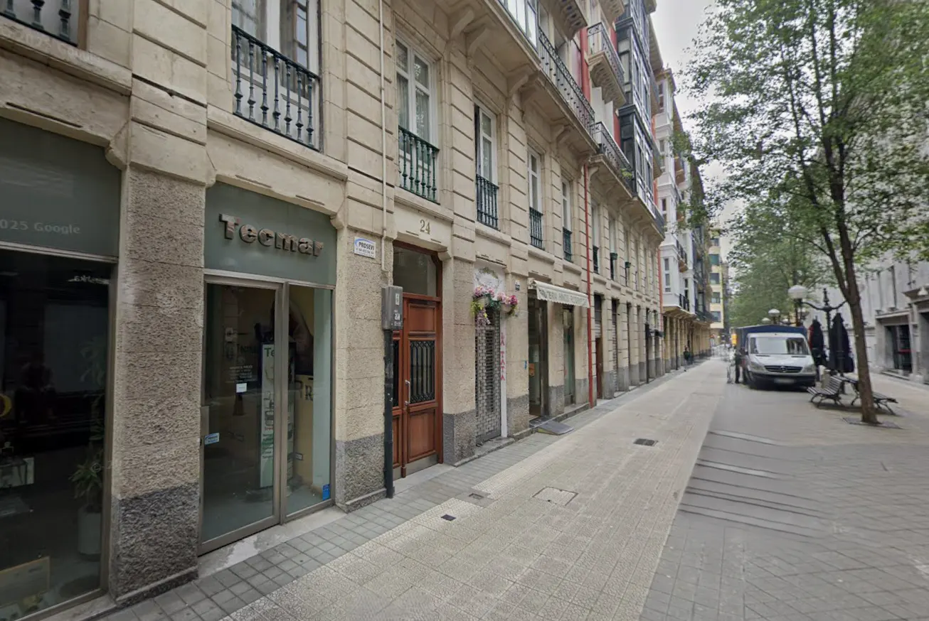 Street view of a row of stone buildings with balconies and shops. A white van is parked on the right side of the street.