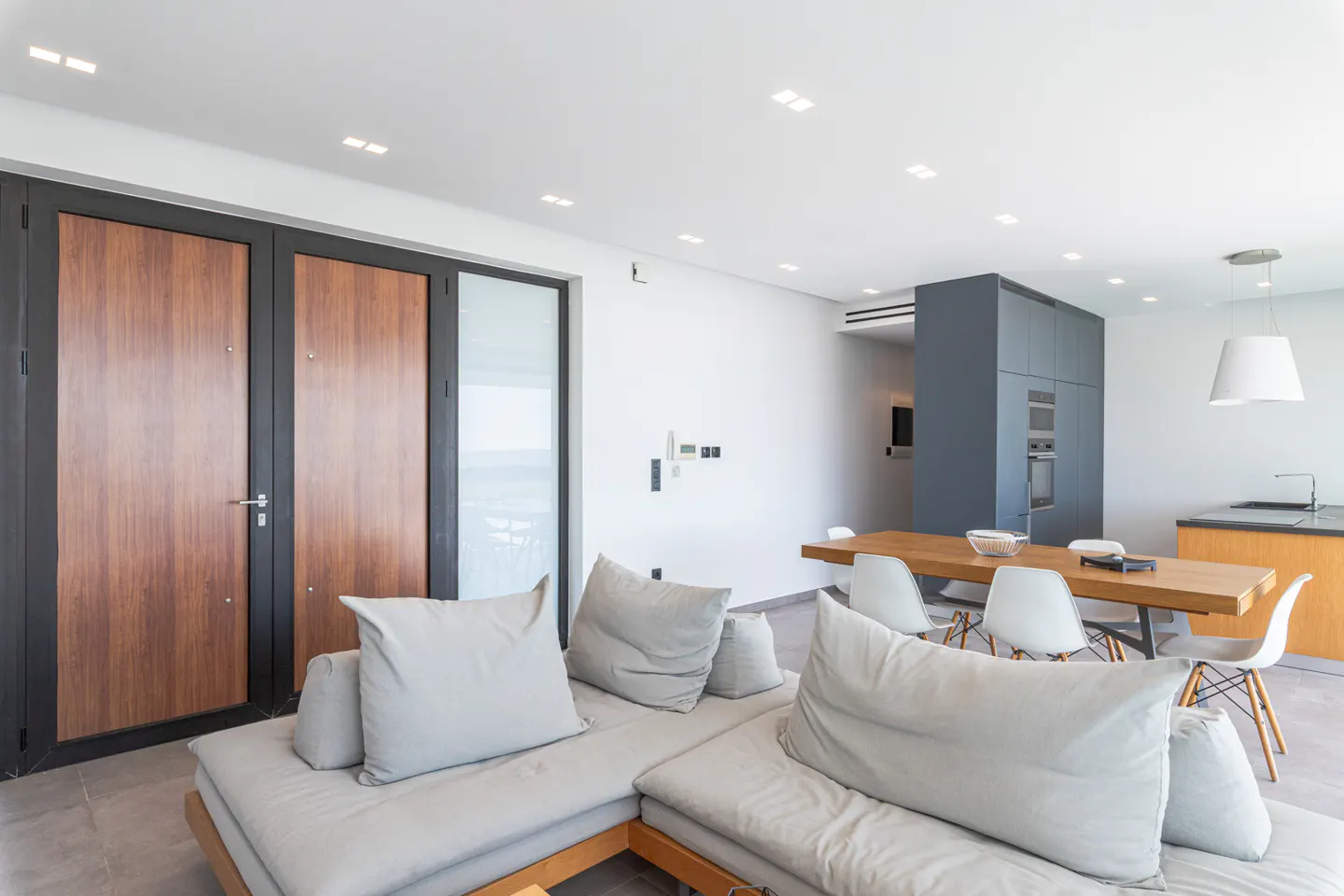 Bright, modern living space with a gray sectional sofa, wood dining table with white chairs, and a gray kitchen with built-in appliances.