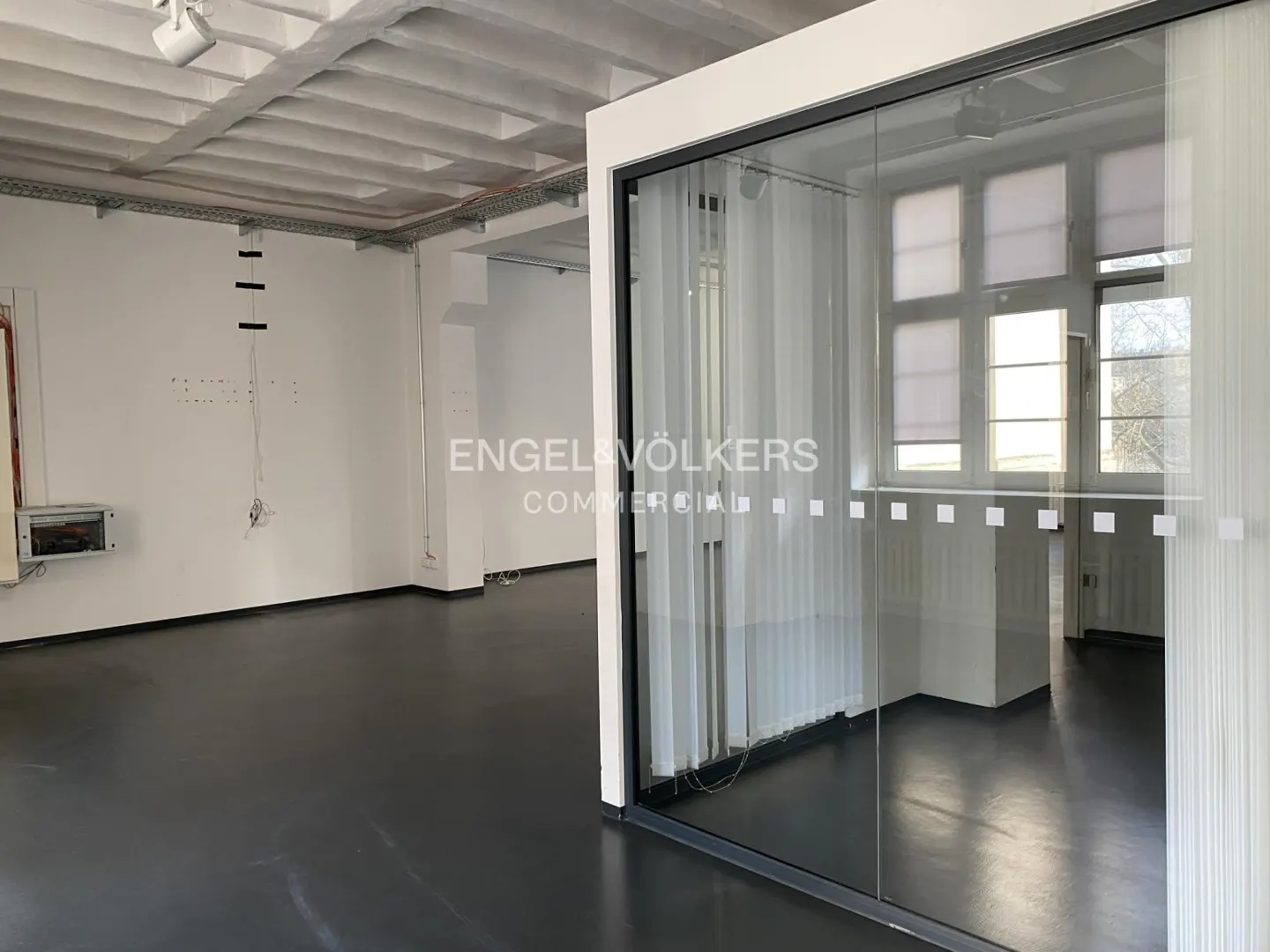 Modern office space with dark floors, white walls, exposed ceiling beams, and a glass-enclosed meeting room.