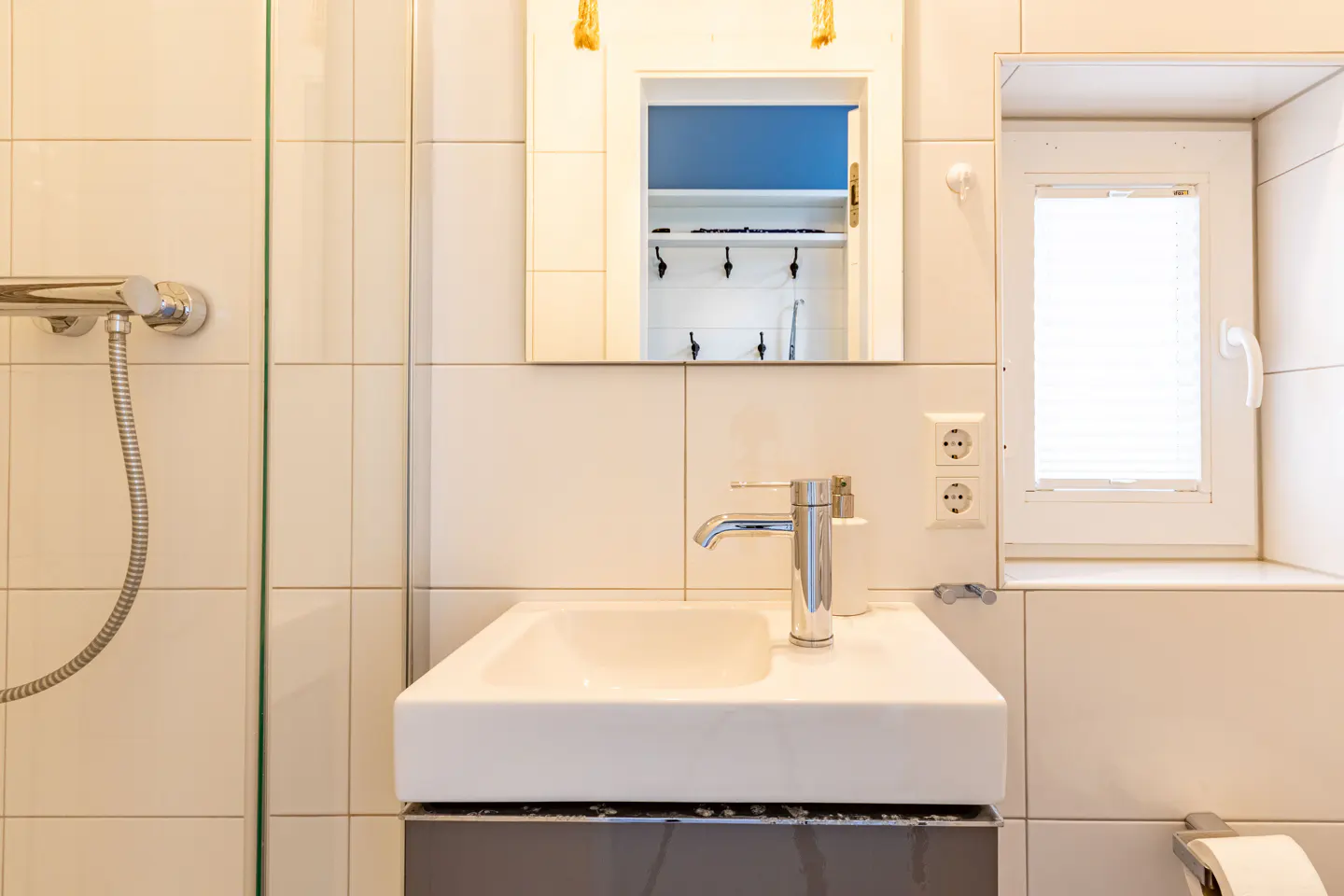 Bathroom with white square tiles, a white sink with a chrome faucet, and a mirror cabinet with hooks inside. A window with blinds is to the right.