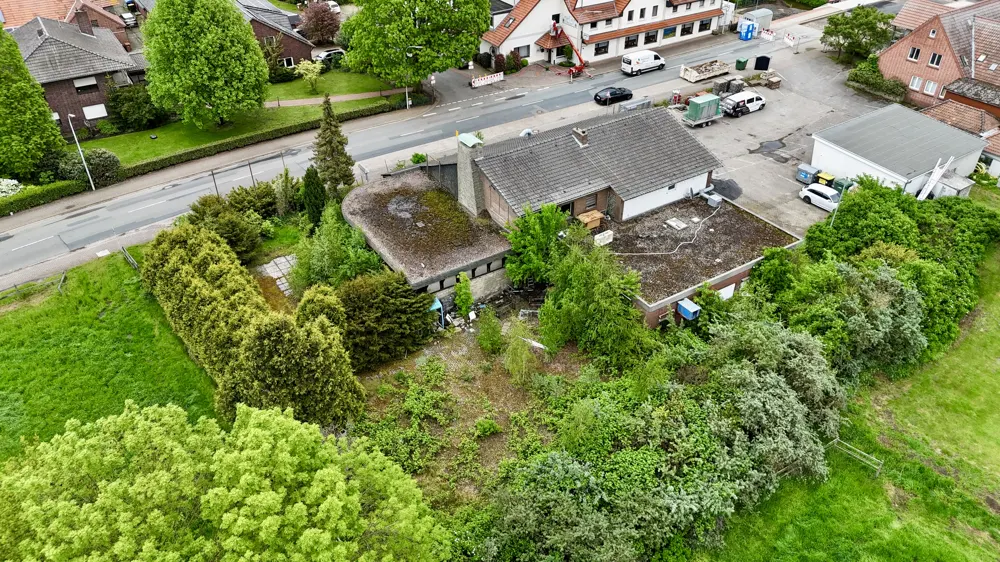 Aerial view of a house with a gray roof, surrounded by green trees and bushes. A road with cars is visible in the background.