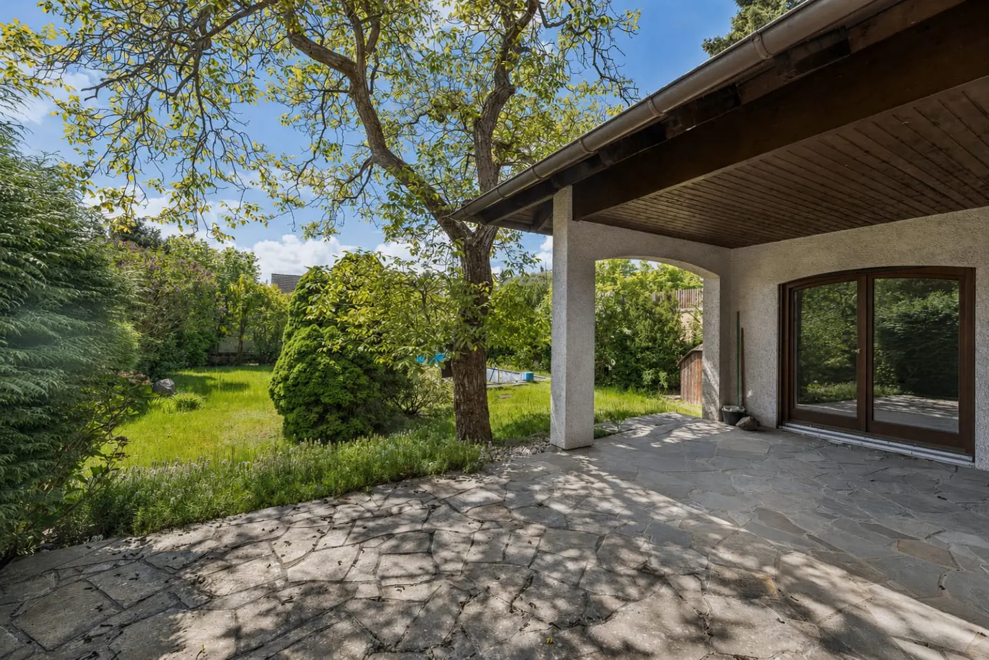 Stone patio with a covered porch, white columns, and brown trim. A green lawn and trees are visible in the background.