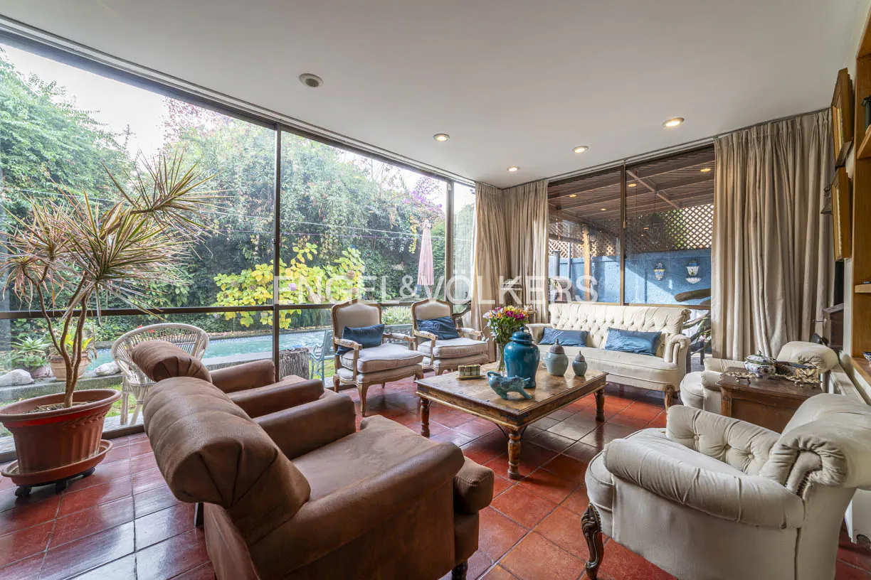 Sunroom with terracotta floors, brown and cream armchairs, a wooden coffee table, and large windows overlooking a pool.