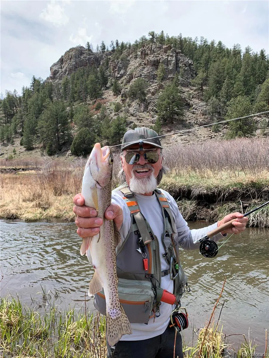 A smiling man in fishing gear holds up a trout by a river, with a mountain backdrop.