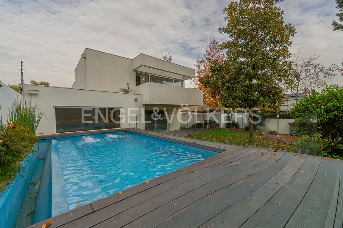 Modern white house with a blue swimming pool and a gray wooden deck in the backyard. Trees and cloudy sky in the background.