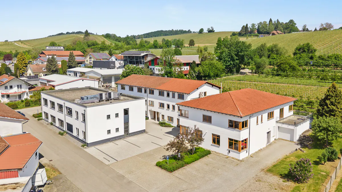 Aerial view of white commercial buildings with red tile roofs, surrounded by a paved parking area and rural green hills.