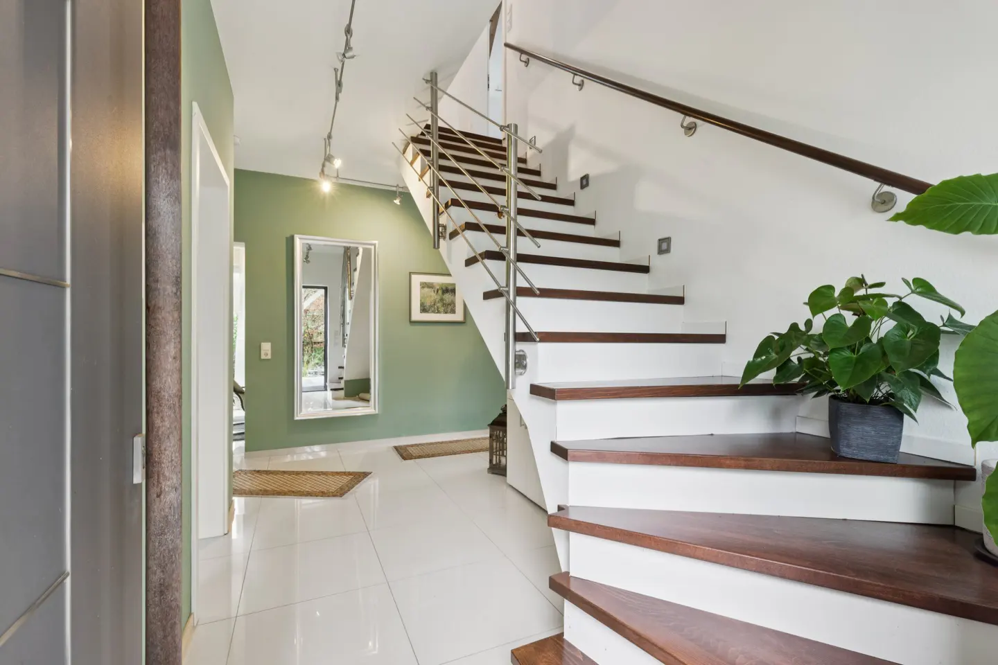 Bright foyer with white tile floor, green wall, and white stairs with wood treads and stainless steel railing. A potted plant sits on the stairs.
