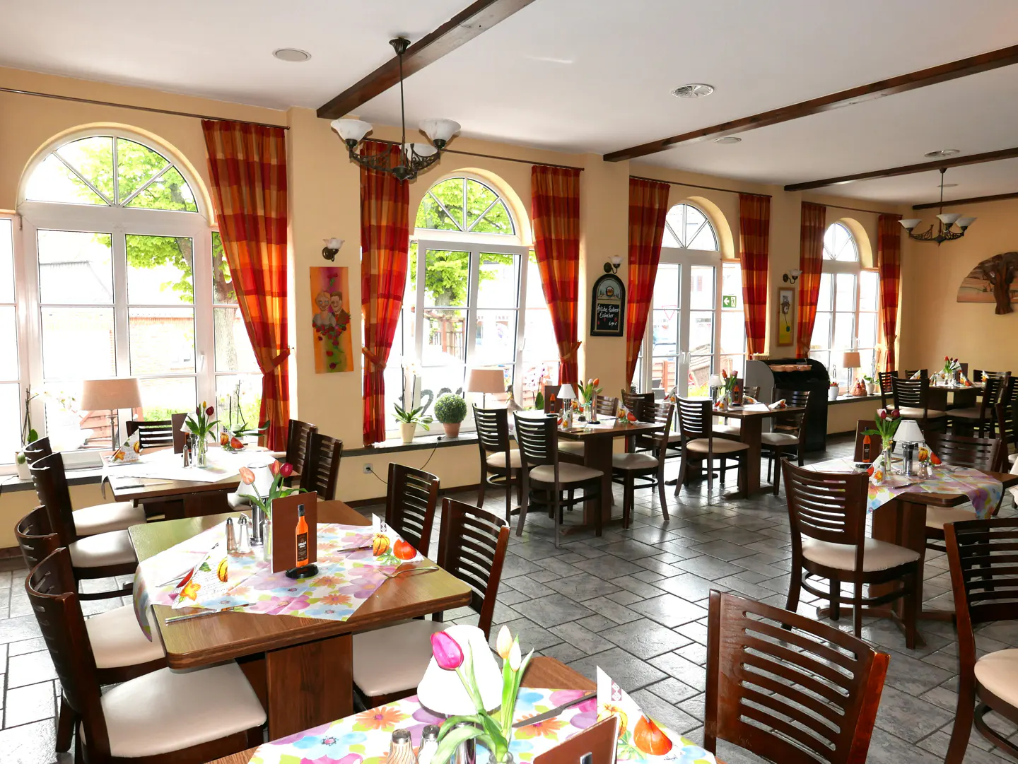 Bright restaurant interior with wooden tables, chairs, floral tablecloths, and large arched windows with orange curtains.