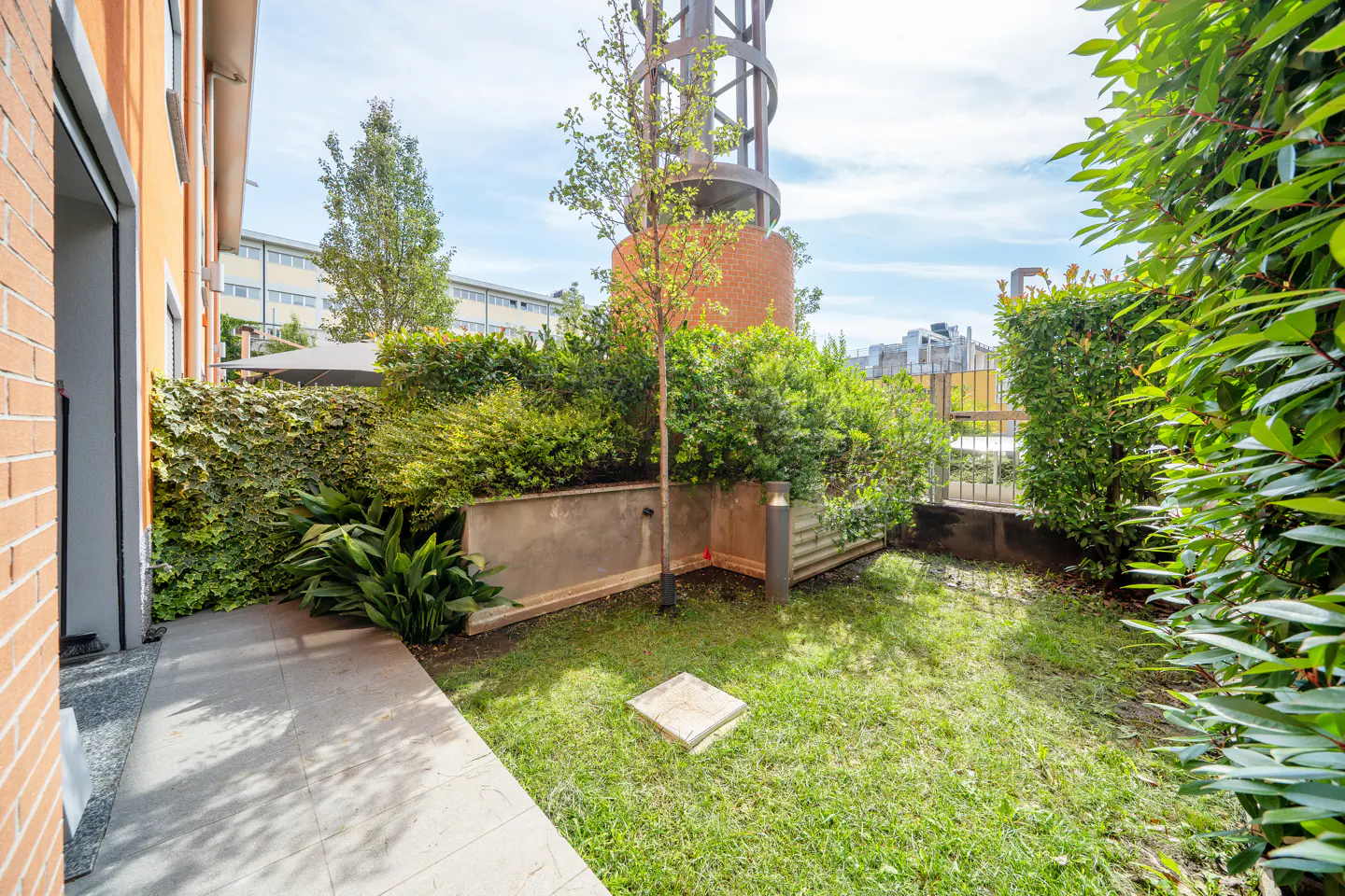 Small private garden with a paved walkway, green lawn, lush shrubs, and a tall brick structure under a bright blue sky.