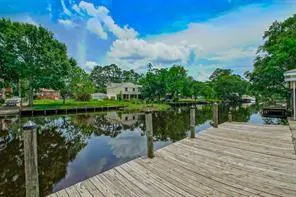 Wooden dock on a canal with trees and houses in the background under a blue sky with white clouds.