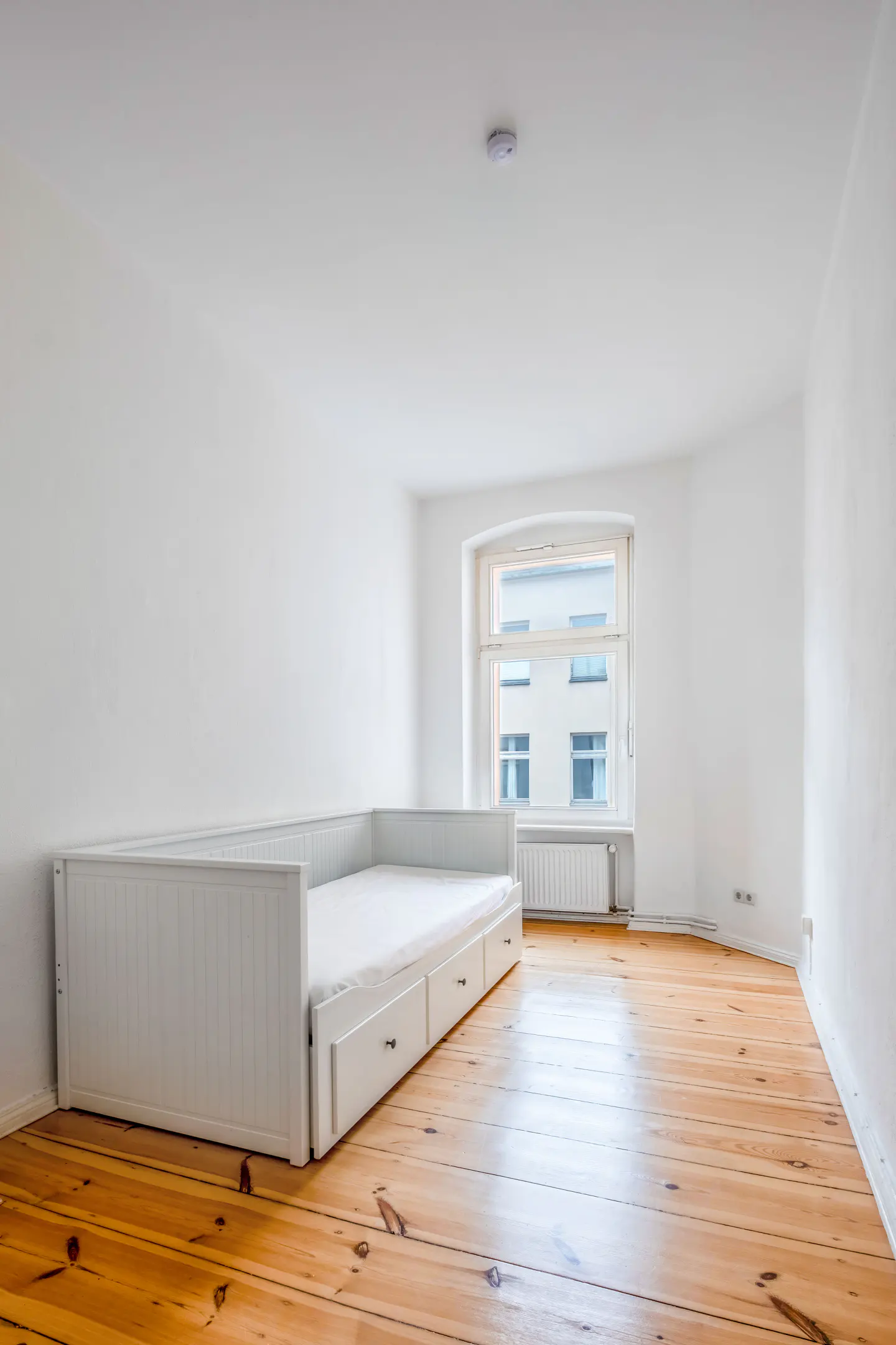 Bright, minimalist bedroom with white walls, a white daybed with storage drawers, and light-toned wooden floorboards.