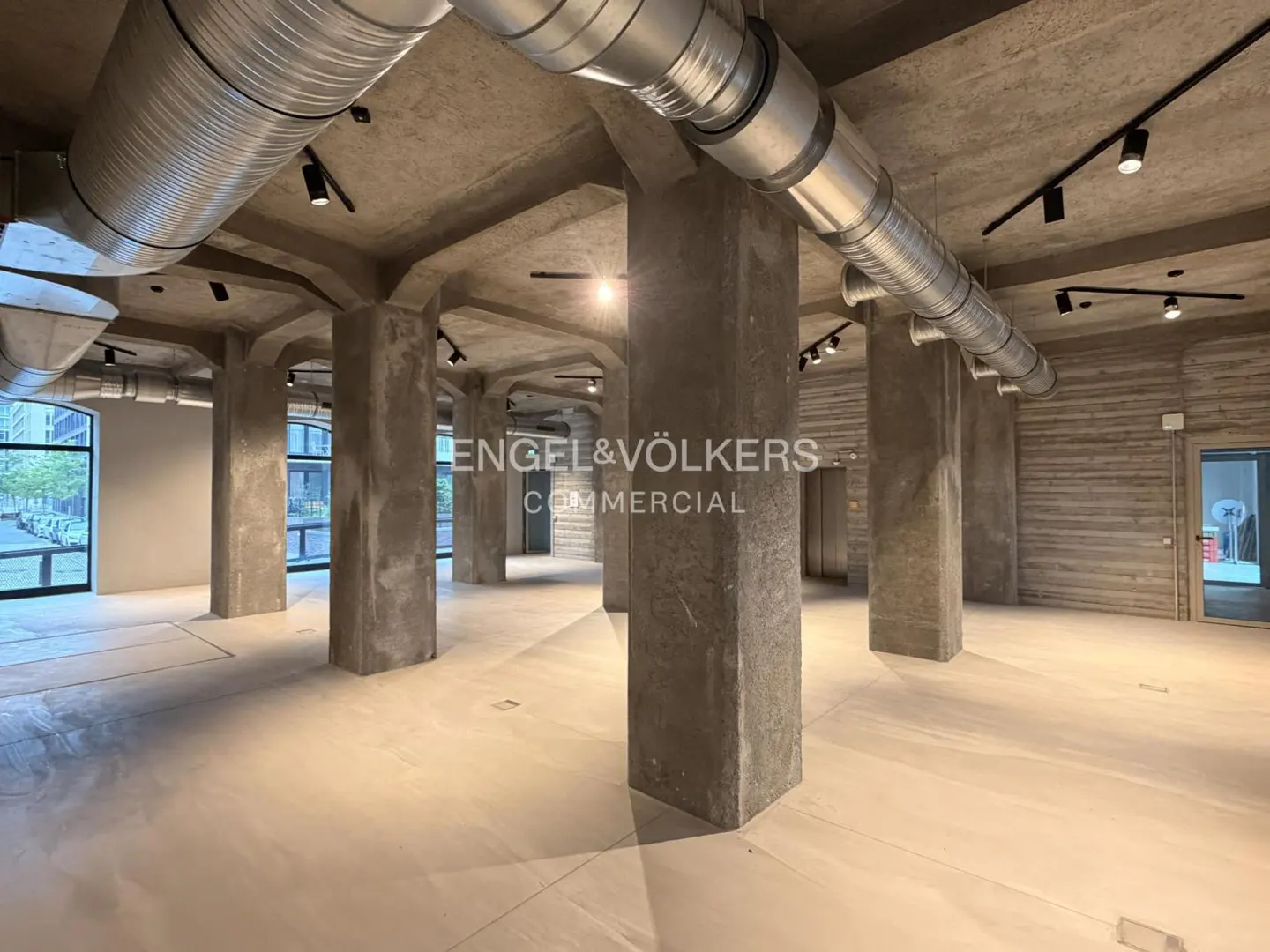 Empty commercial space with concrete pillars, floors, and ceiling. Silver ductwork and black track lighting run across the ceiling. Engel & Völkers logo is visible.