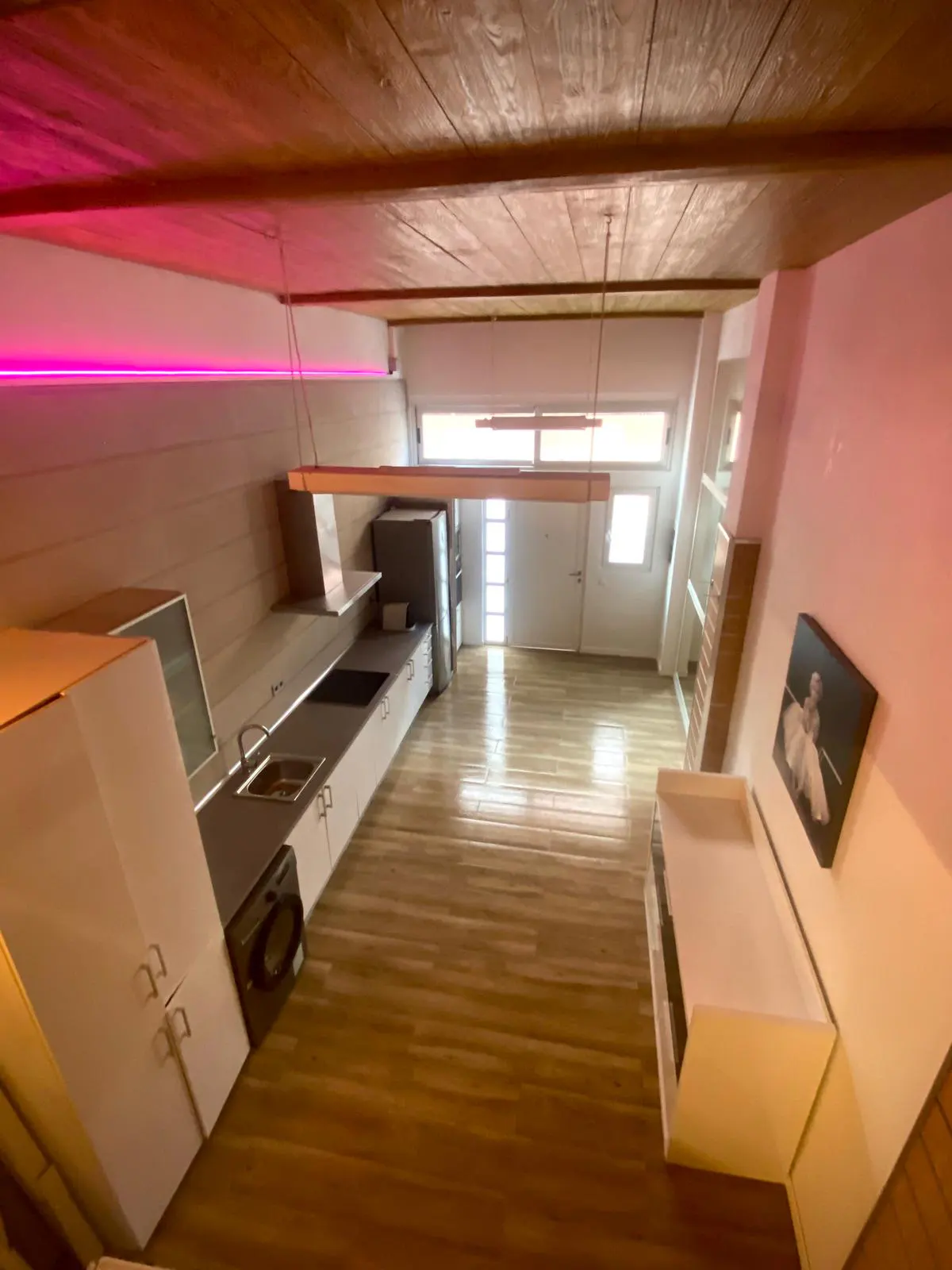 High-angle view of a modern kitchen with wood ceilings, pink accent lighting, white cabinets, and light wood flooring.