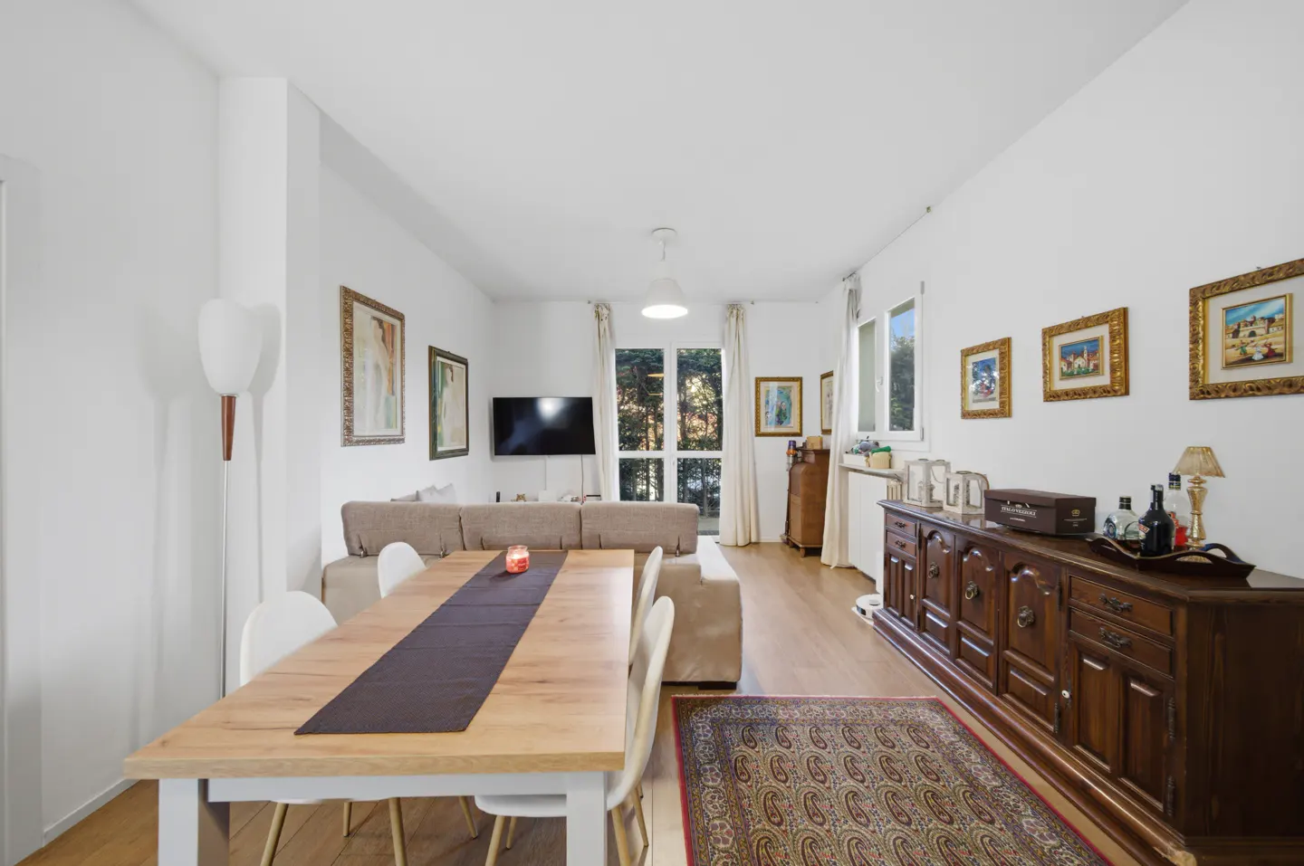 Bright living room with a wooden dining table, beige sofa, and dark wood cabinet. Art hangs on the white walls.