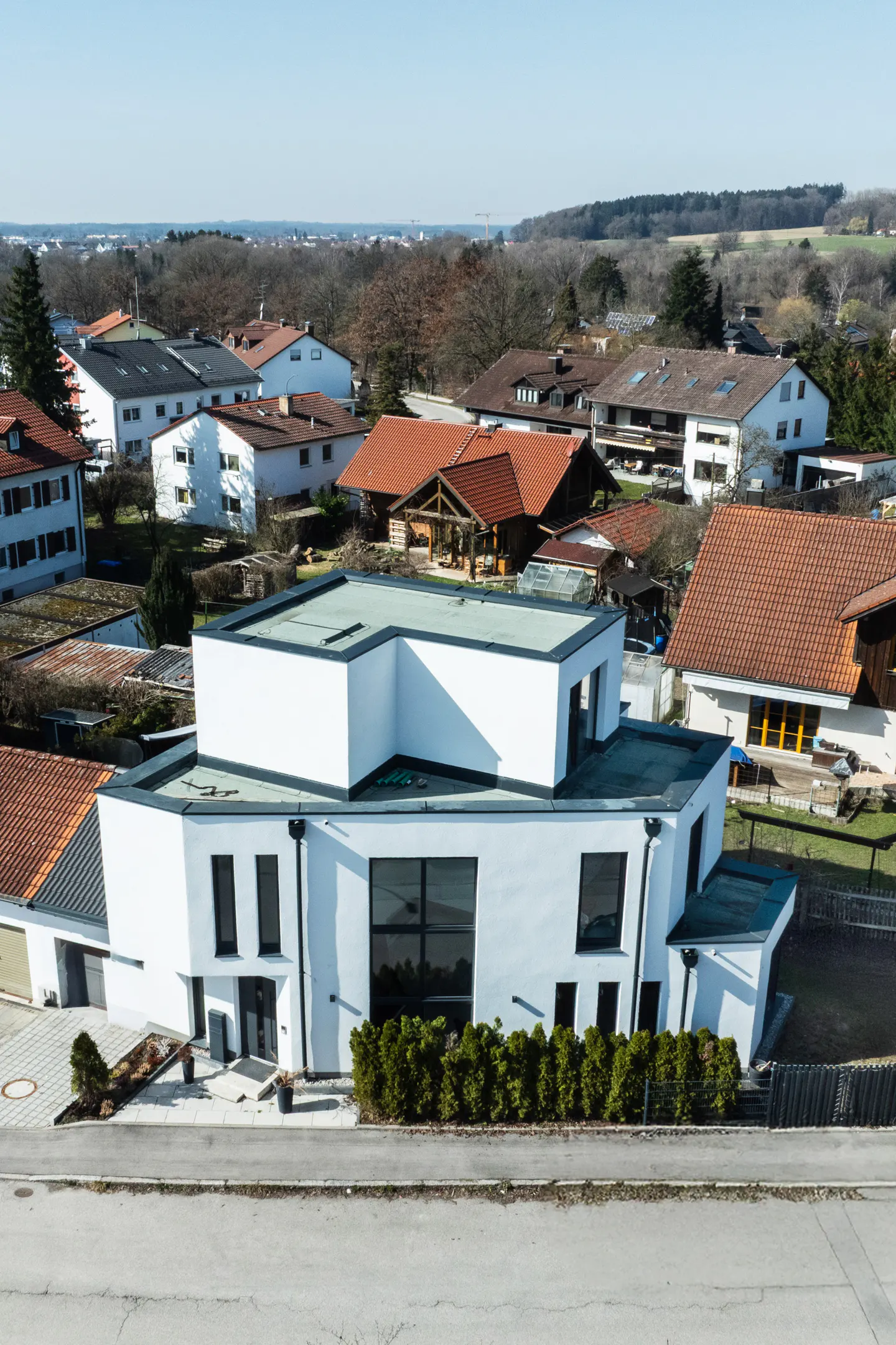 A modern, white, two-story house with a flat roof, surrounded by other houses and trees.