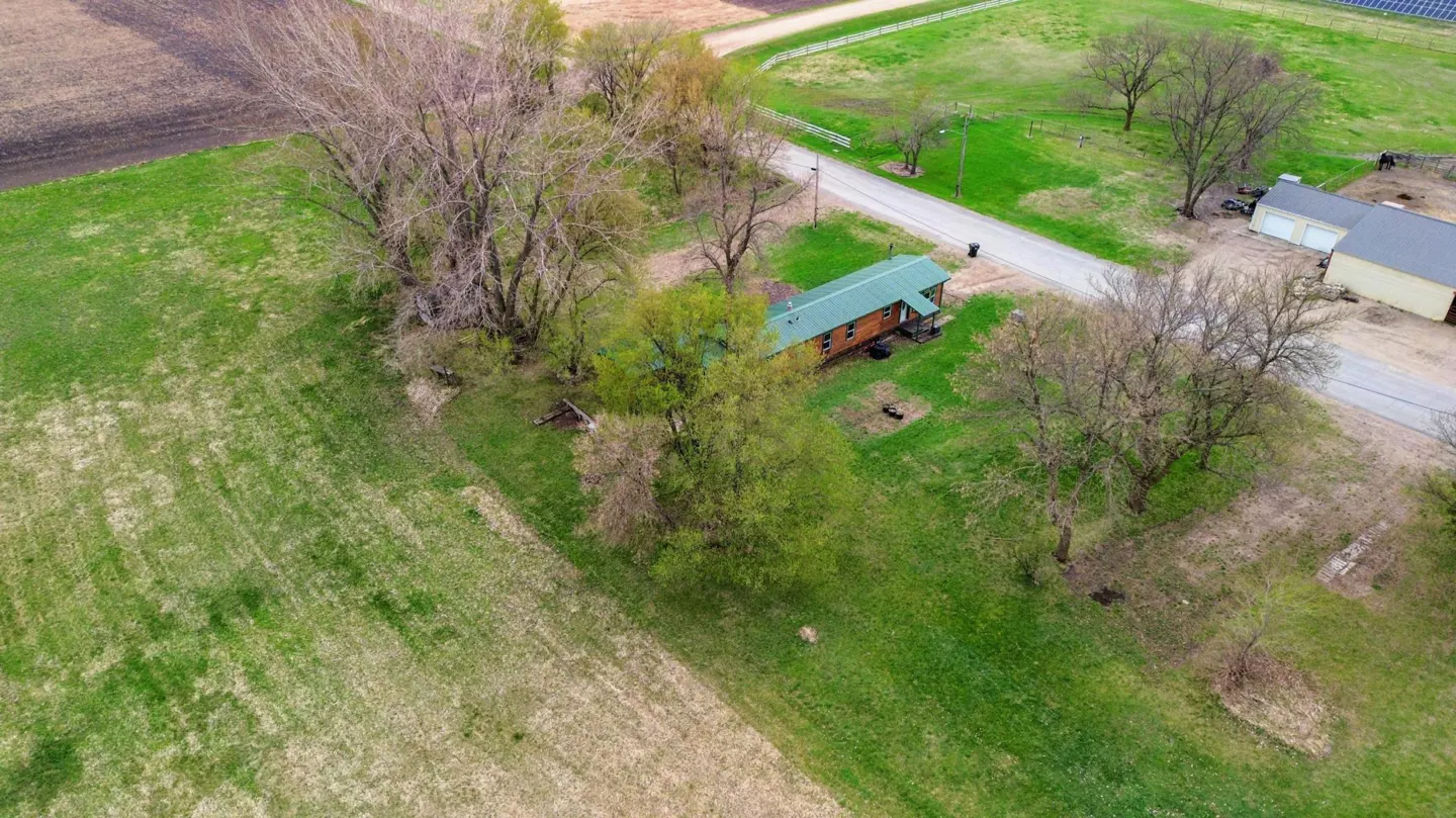 Aerial view of a brown house with a green roof, surrounded by green grass, trees, and farmland. A road runs alongside the property.