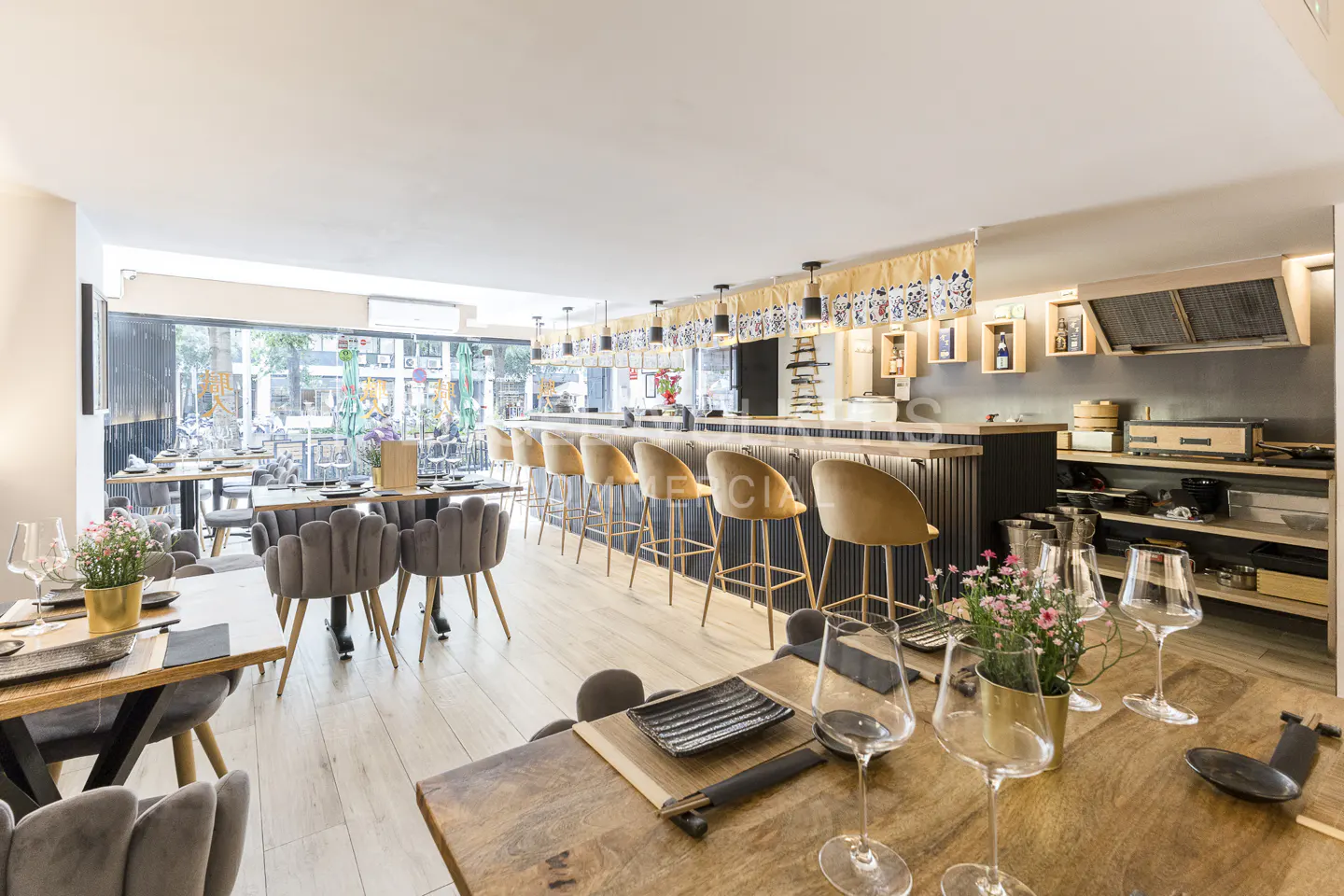 Upscale restaurant interior with wooden tables, grey chairs, and a bar with tan stools. Large windows let in natural light.