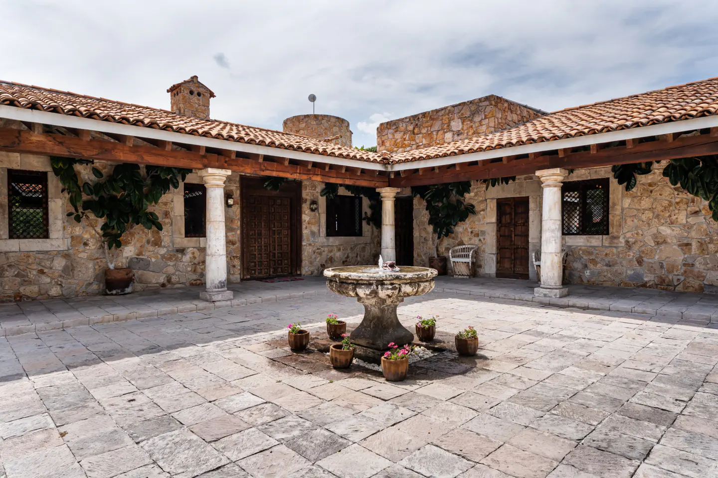 Courtyard view of a stone house with a fountain in the center, surrounded by potted plants with pink flowers.