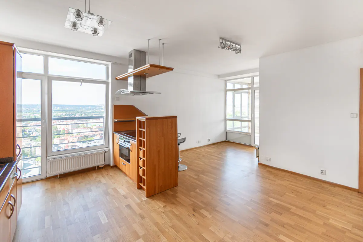 Bright apartment interior with wood floors, white walls, and a kitchen area with wood cabinets and a large window overlooking a city.