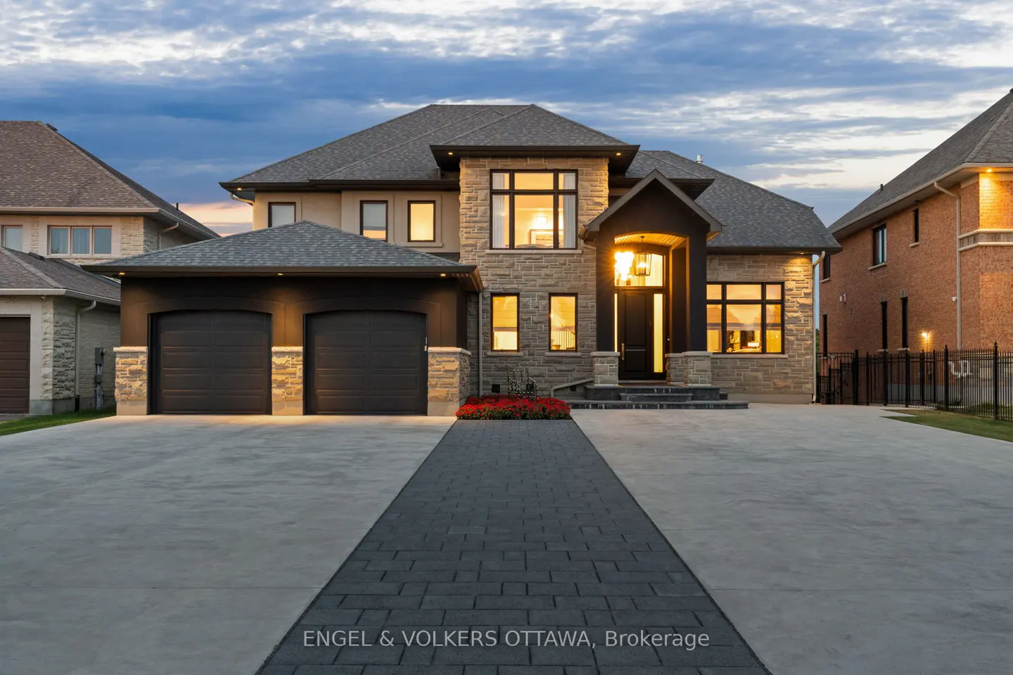Exterior view of a two-story stone house with a gray roof, black garage doors, and a paved driveway. Red flowers accent the front entrance.