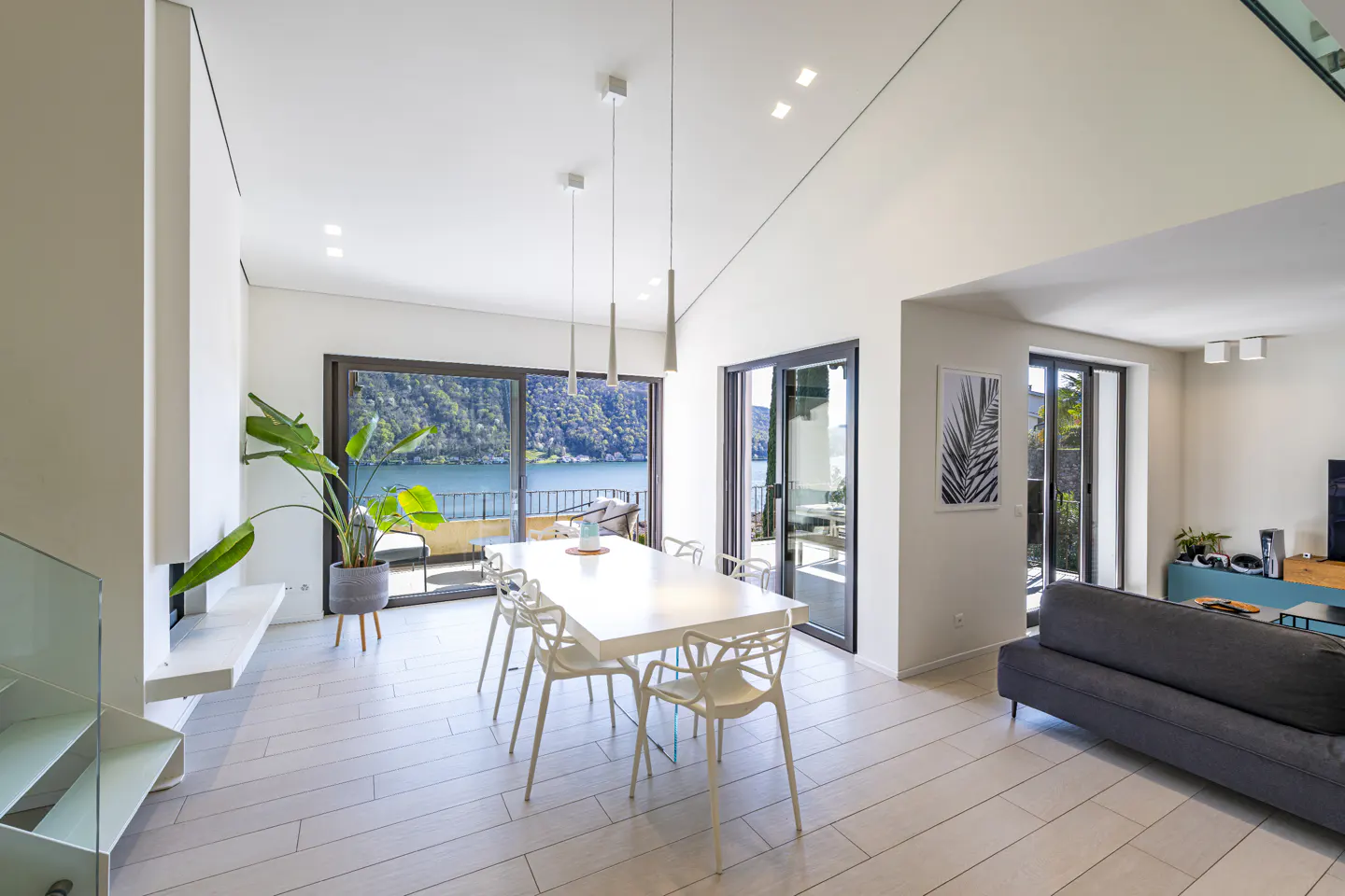Bright, modern dining room with a white table, chairs, and wood floors. Large windows offer a scenic lake view. A gray sofa is visible in the adjacent living area.