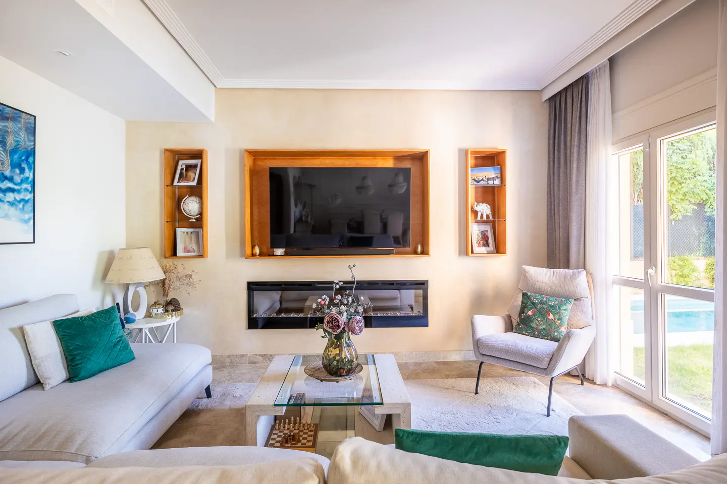 Living room with a TV over a fireplace, a white sofa with green pillows, and a glass coffee table. Natural light from a window.