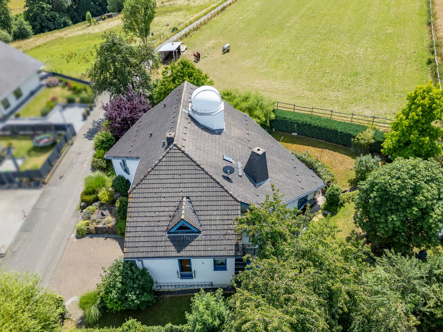 Aerial view of a white house with a gray tiled roof, a white dome, and a chimney, surrounded by green trees and a field with horses.
